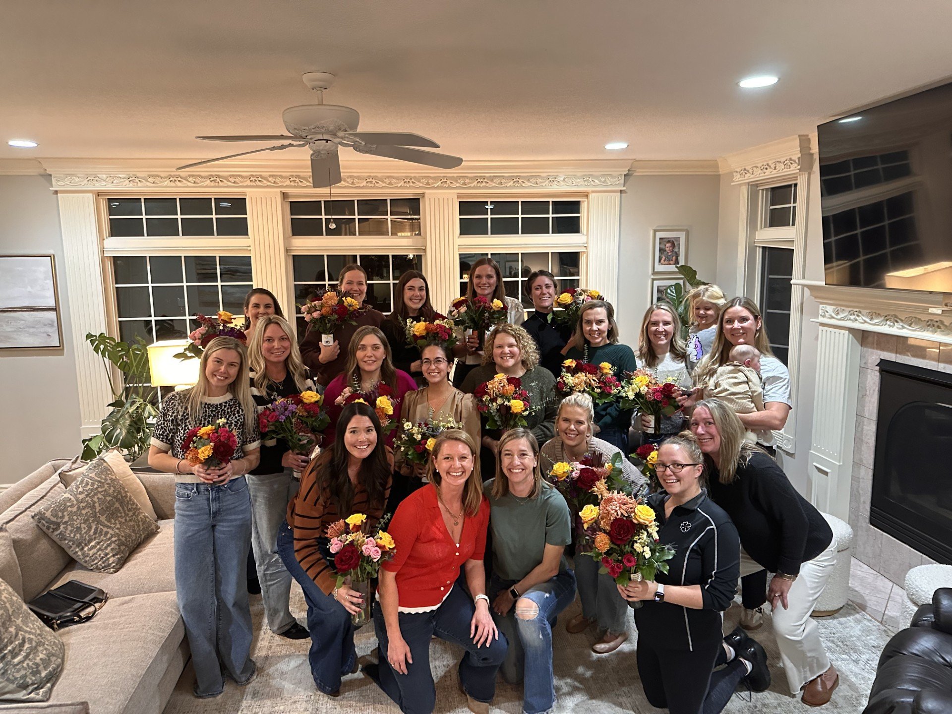 Group of women gathered indoors holding bouquets of flowers, smiling for a photo in a warmly decorated living room with large windows, a fireplace, and various decorations.