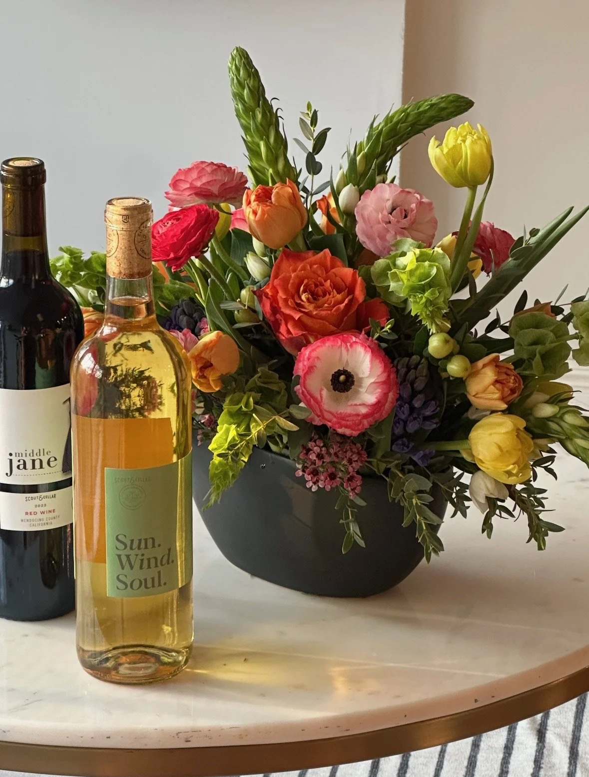 A bouquet of colorful flowers in a black vase, two bottles of wine, and a white marble table with a striped cloth underneath.
