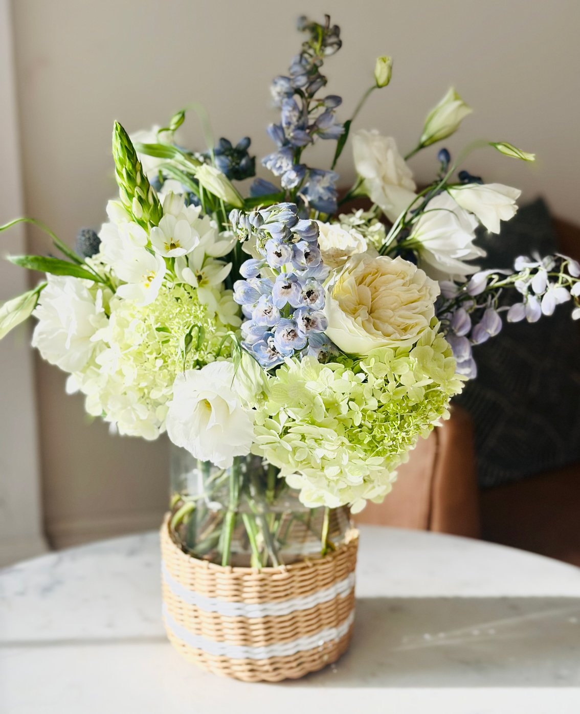 A bouquet of white and light purple flowers in a glass vase with a woven basket around its base, placed on a white marble table.