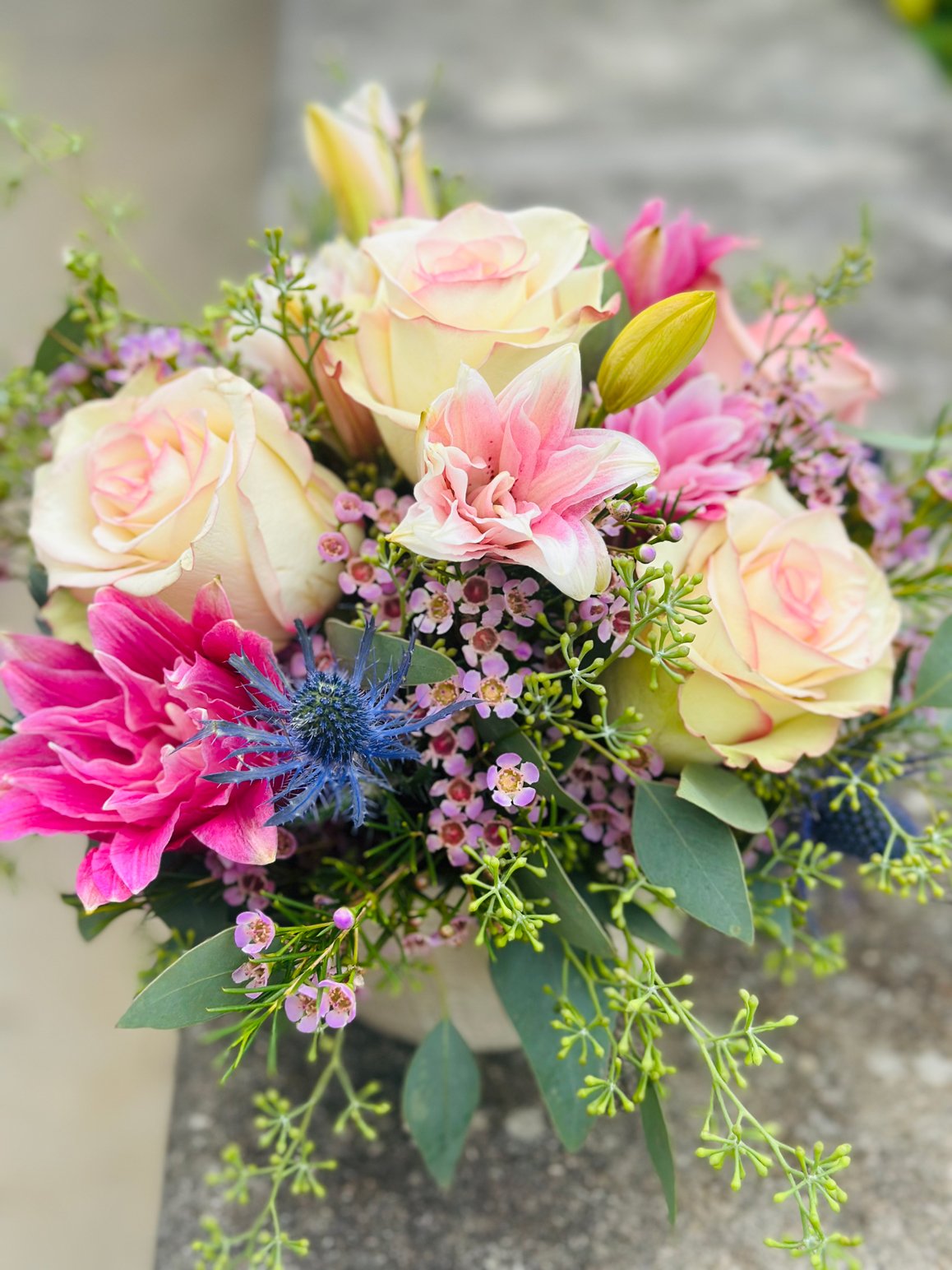 A colorful bouquet of various flowers including pale pink roses, pink lilies, purple thistle, and small pink and purple filler flowers, with green foliage, placed outdoors on a stone surface.
