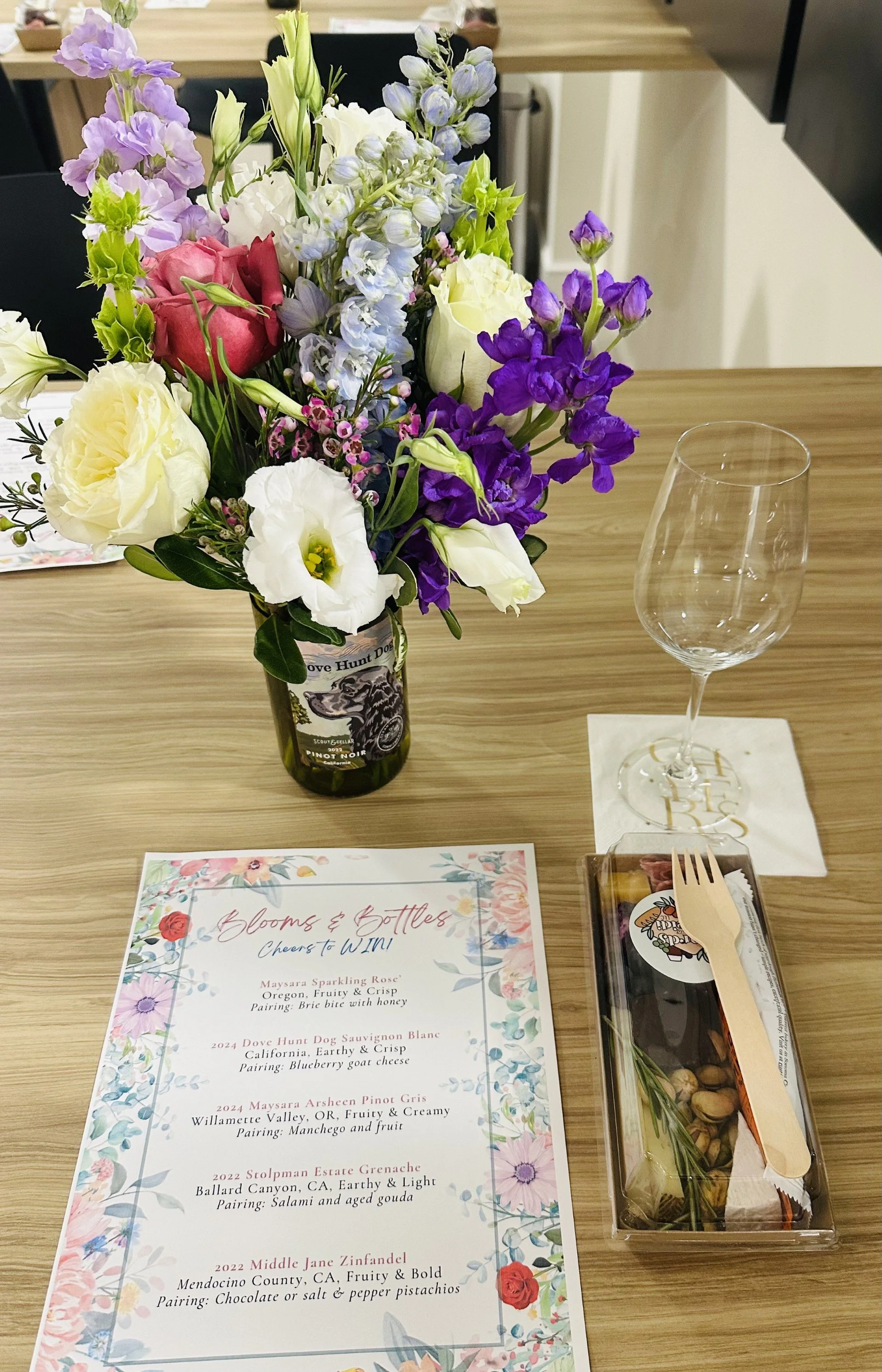 A floral arrangement in a wine bottle vase placed on a wooden table, with a wine glass on a napkin, a menu, and a snack box nearby.