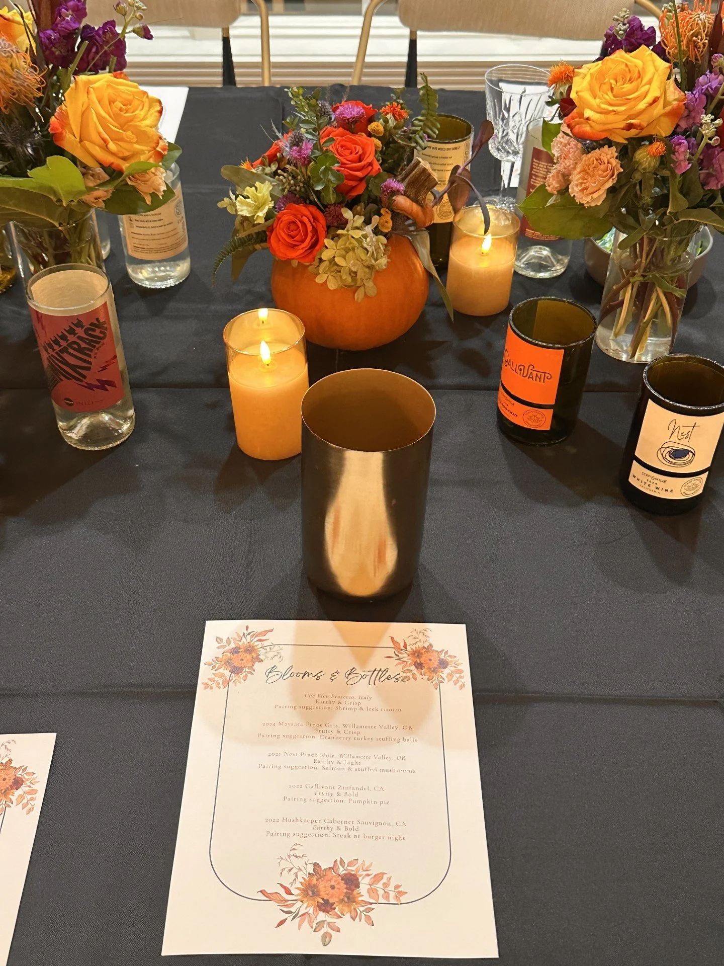 A table decorated with fall-themed floral arrangements, candles, and wine bottles, with a menu titled 'Blooms & Bottles' featuring wine and food pairings.