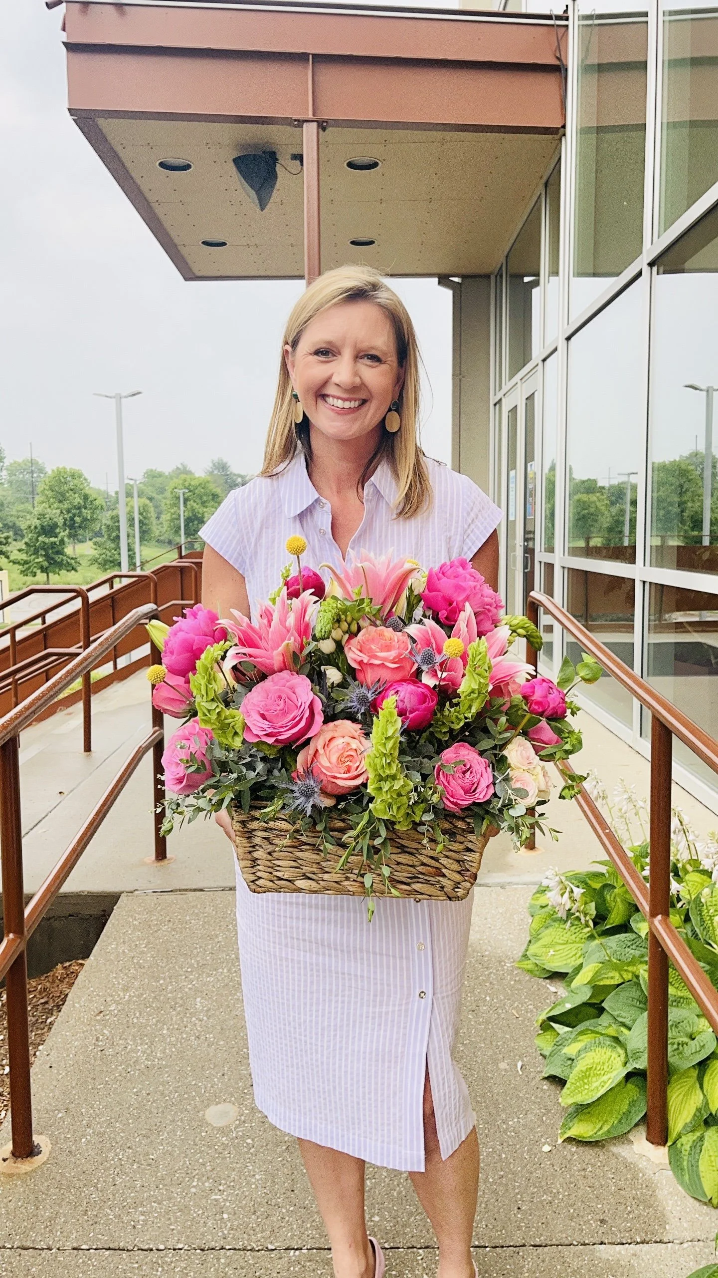 A woman with blonde hair wearing a light purple striped dress stands outside of a building with glass windows, holding a large basket of pink and peach flowers.