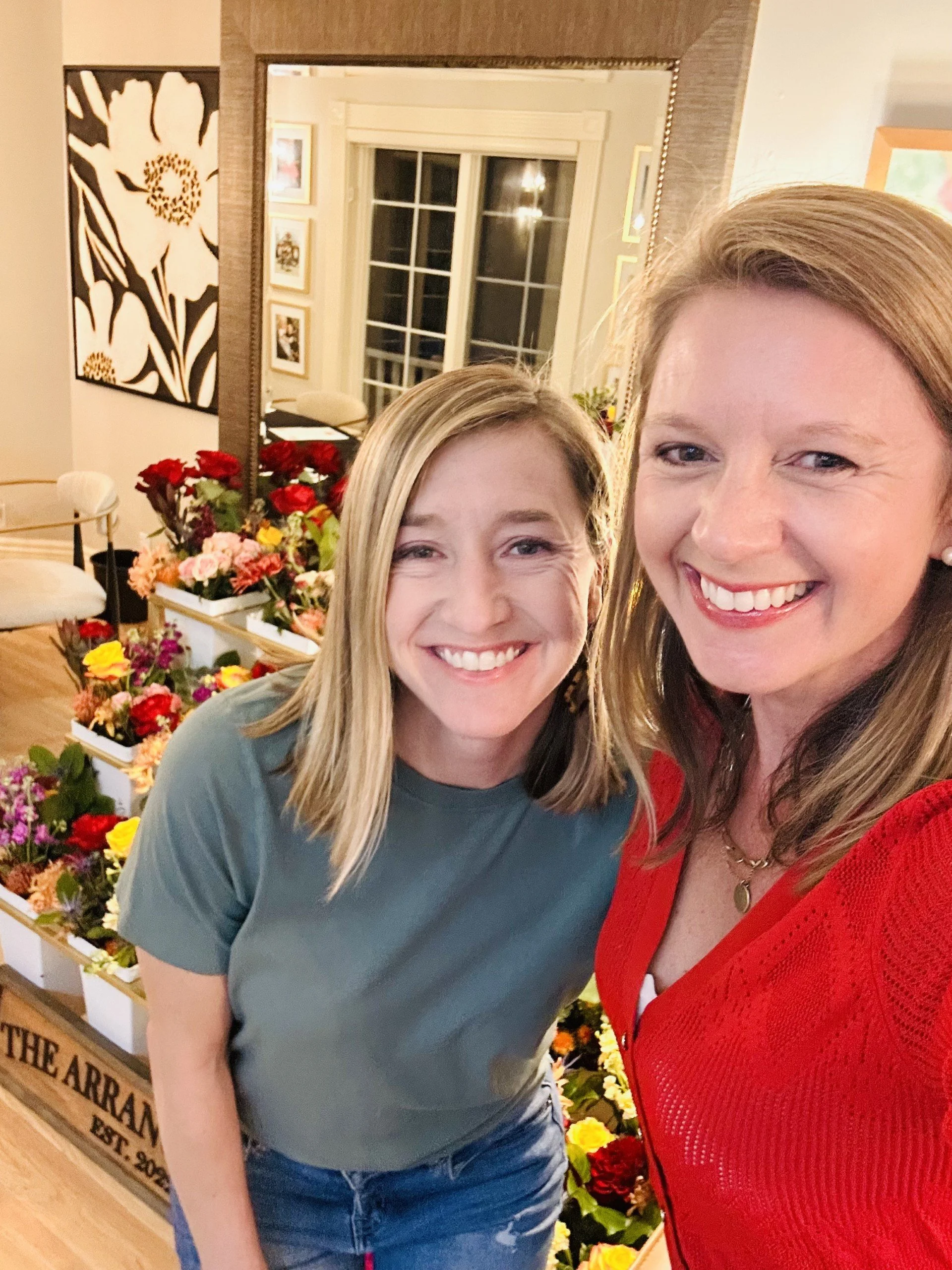 Two women smiling for a selfie in front of a large mirror with a display of colorful flowers behind them.