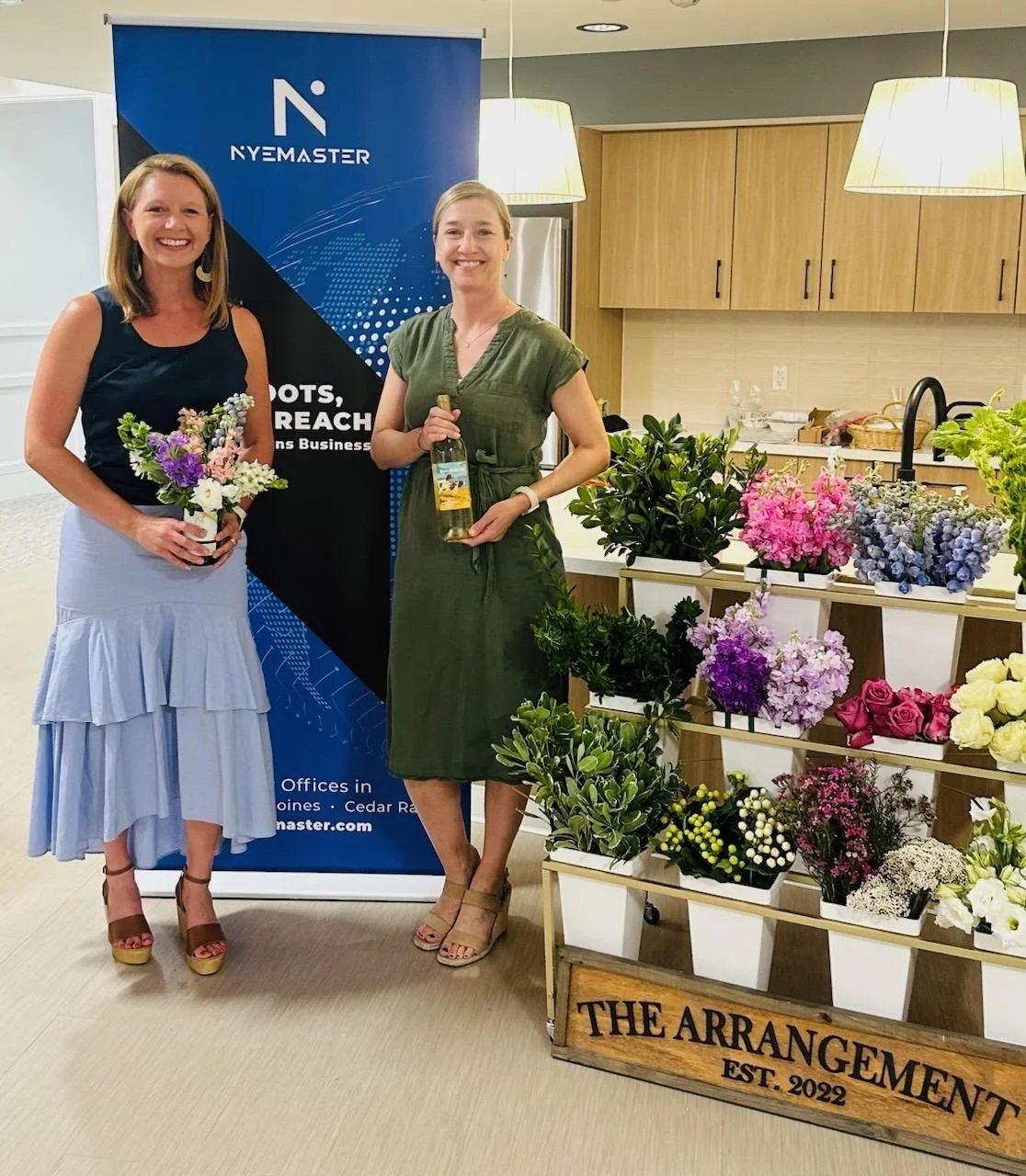 Two women standing indoors next to a flower display and a banner for NYE Master, with one holding a bouquet and the other holding a bottle of wine. Both women are smiling.