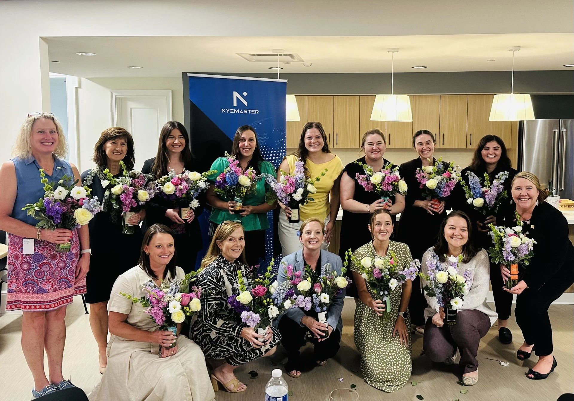 Group of women indoors, smiling and holding floral bouquets, with a banner that says 'NYEMASTER' in the background, beige cabinets, and pendant lights above.