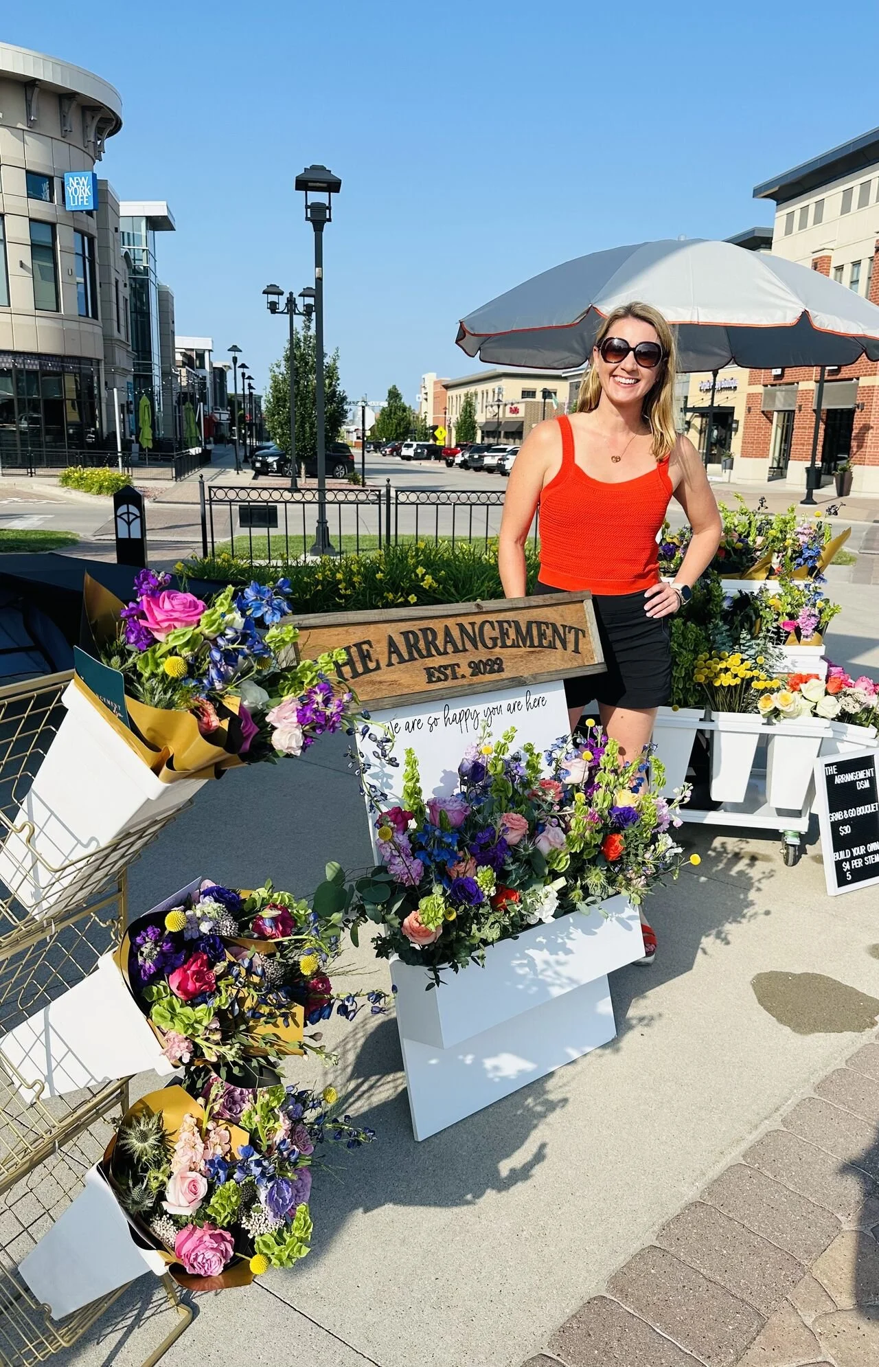 A woman with blonde hair wearing sunglasses, an orange top, and black shorts standing next to a flower stand with a sign that reads 'The Arrangement EST. 2022'. The stand is decorated with colorful flowers and a smaller sign that says 'We are so happy you are here'. There are city buildings, cars, and a blue sky in the background.