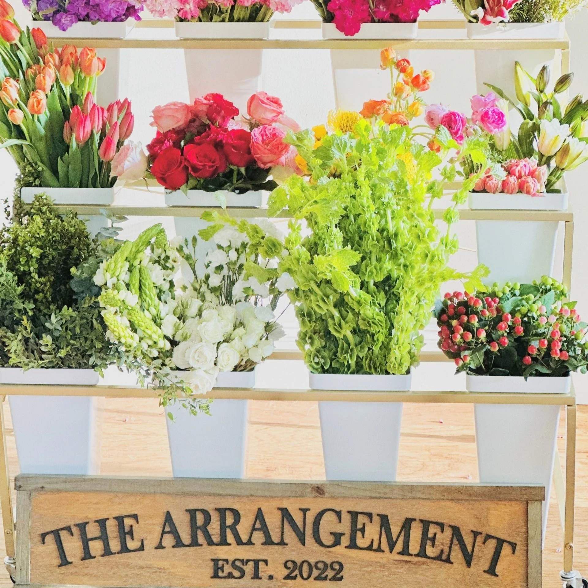 Display of various colorful flowers arranged in white pots on a stand with a wooden sign reading 'The Arrangement Est. 2022' in front.
