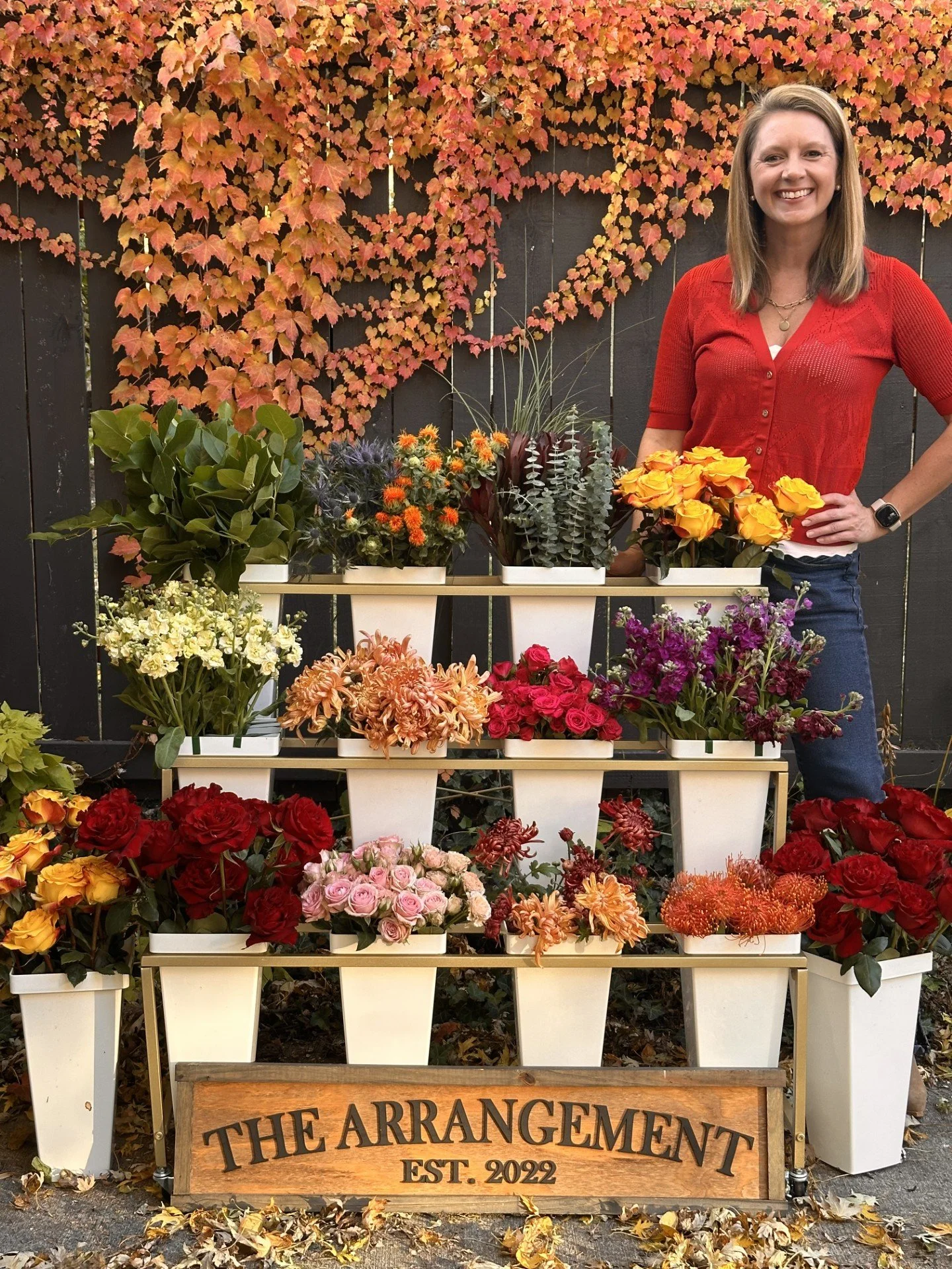 A woman with shoulder-length brown hair, wearing a red cardigan and dark jeans, stands next to a display of colorful flowers in white pots. The flowers include yellow, pink, red, and orange blooms, arranged on a tiered white stand, with a backdrop of
