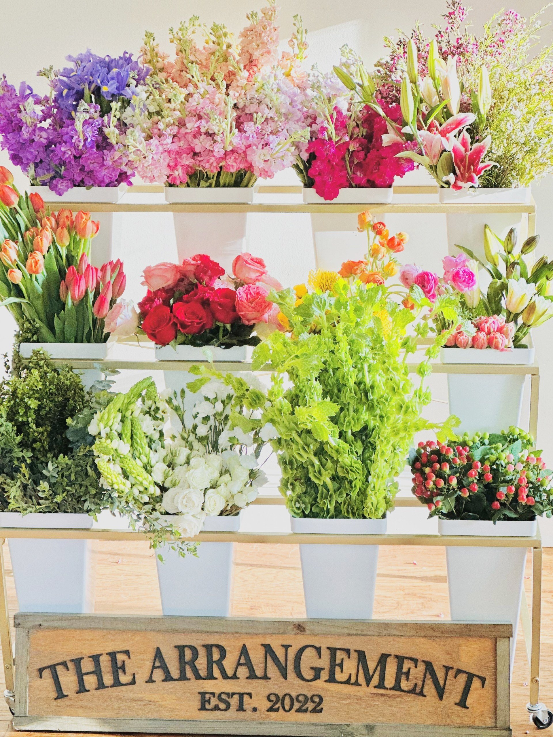 Display of colorful flower arrangements on a multi-tiered stand with a sign reading 'The Arrangement Est. 2022' in front.