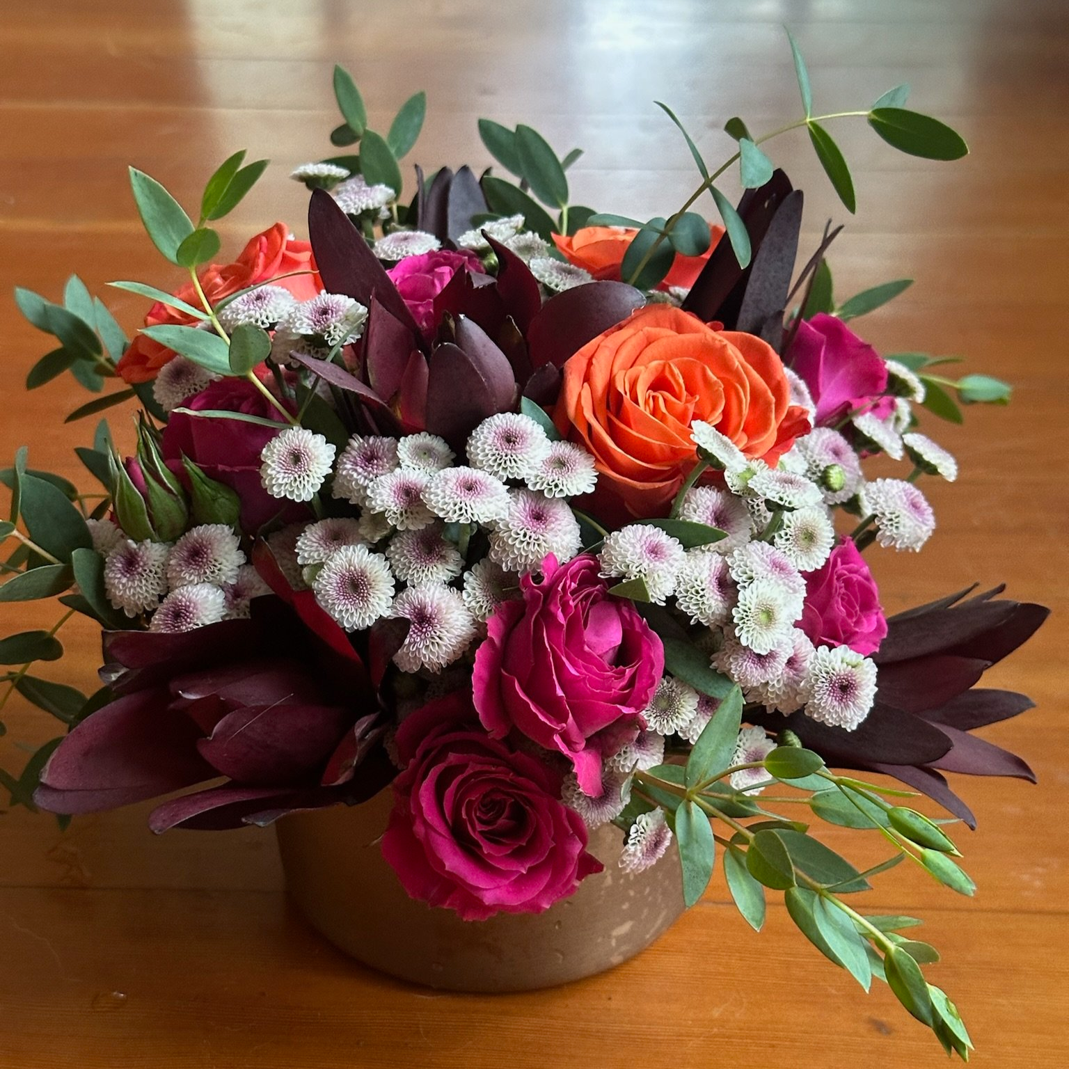 A bouquet of orange, pink, and purple roses with white small daisy-like flowers and green leaves in a brown vase on a wooden surface.
