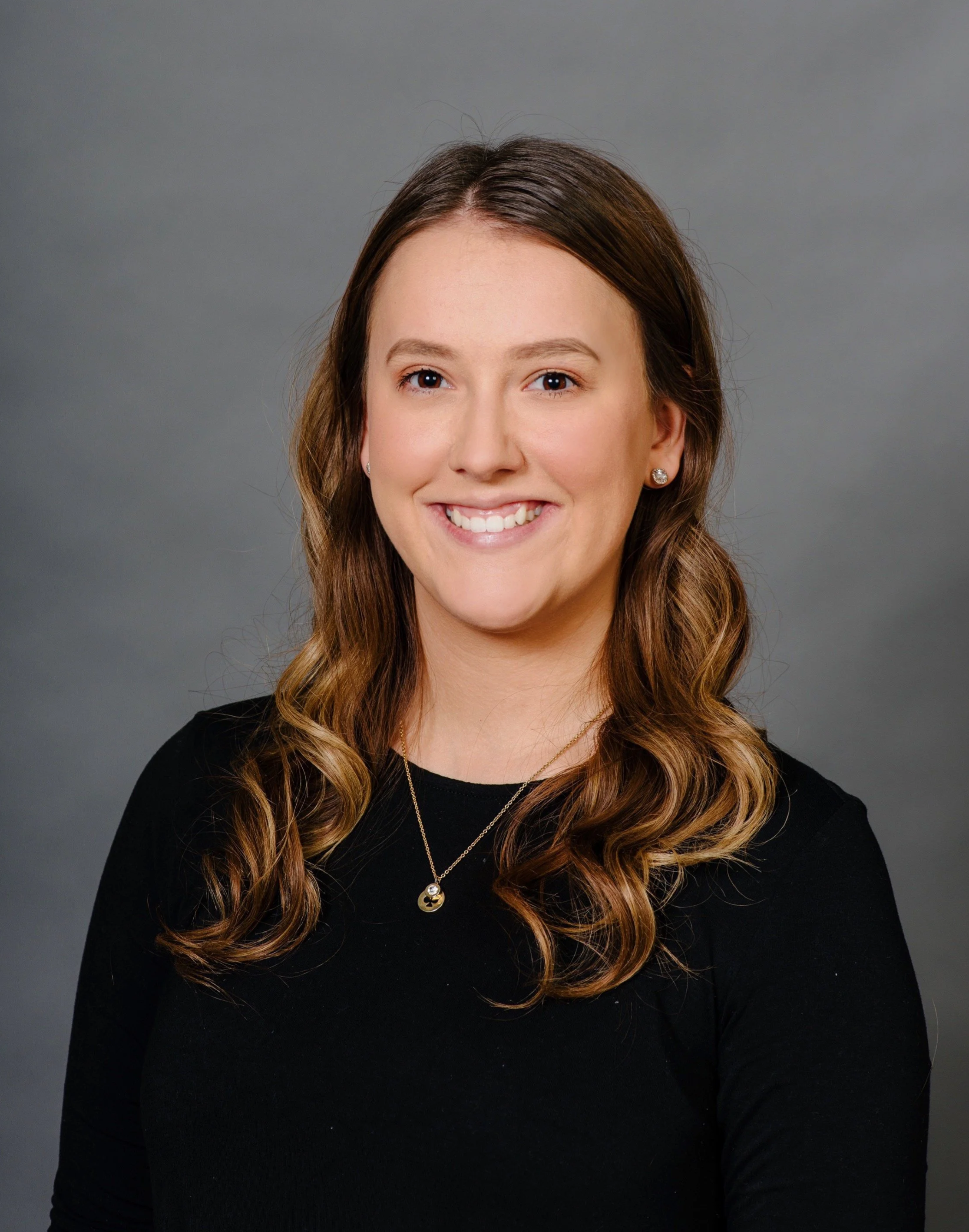 A young woman with long brown wavy hair wearing a black top and jewelry, smiling in front of a plain gray background.