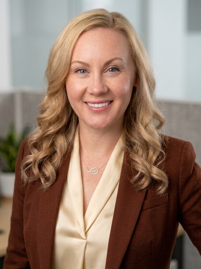 A woman with blonde hair smiling, dressed in a brown blazer over a cream-colored blouse, wearing a silver necklace with a swirl pendant, in an office setting.