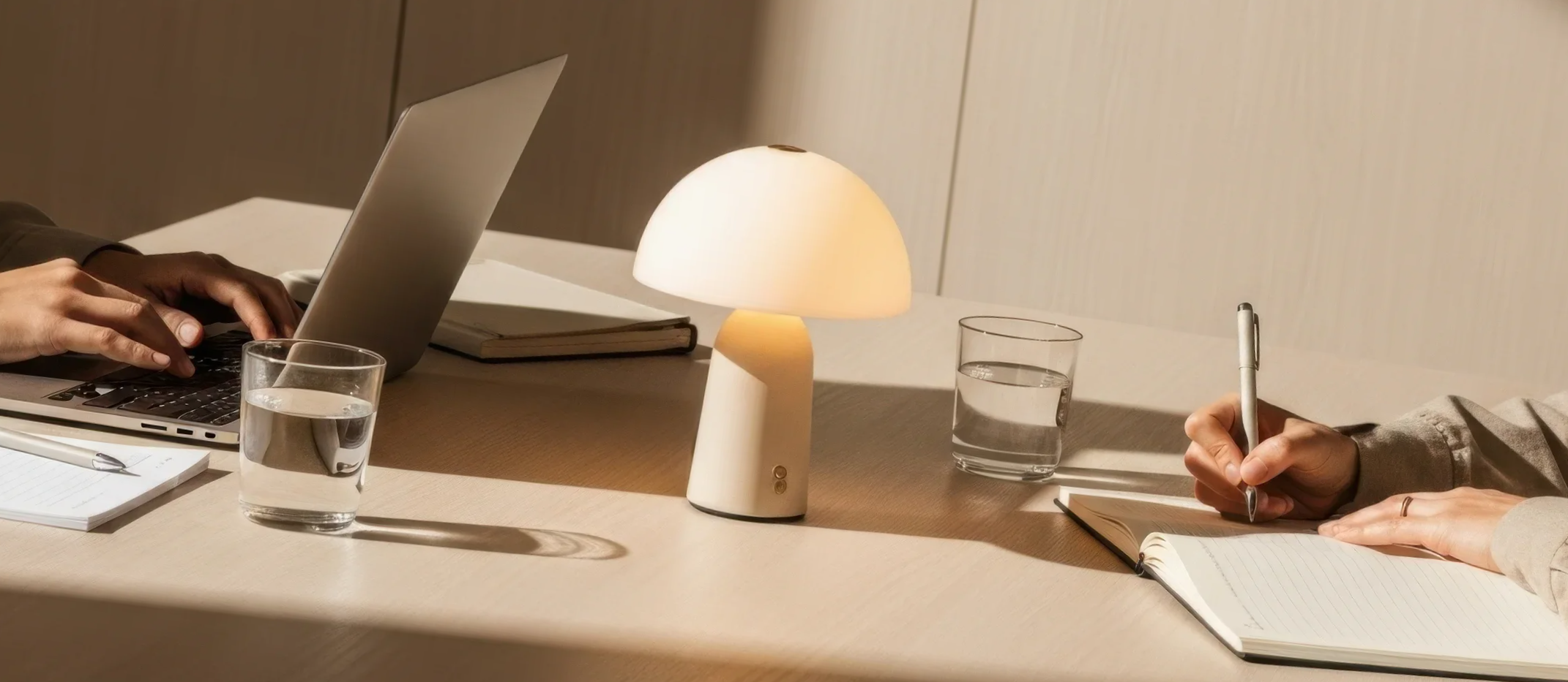 Two people working at a beige table with a lamp, notebooks, glasses of water, and a laptop.