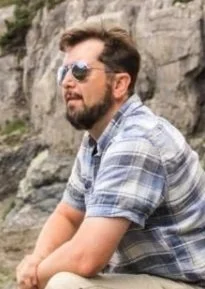 A man with a beard and sunglasses sitting outdoors near rocks, wearing a short-sleeve plaid shirt.
