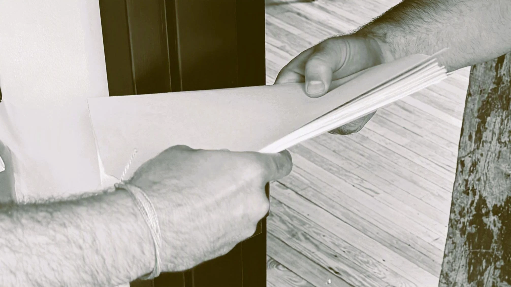 Person holding a book or magazine, showing the page to another person. The scene is indoors, with a wood floor and a dark cabinet or door in the background.