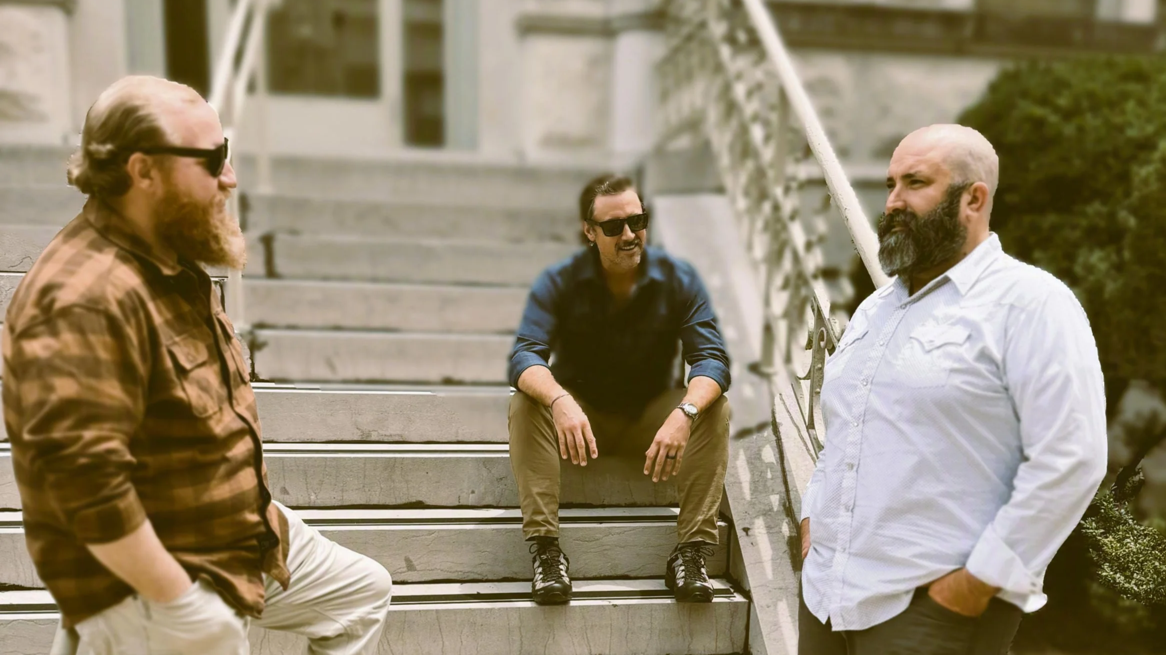 Four men with beards and sunglasses talking and relaxing on the steps outside a building, with greenery nearby.