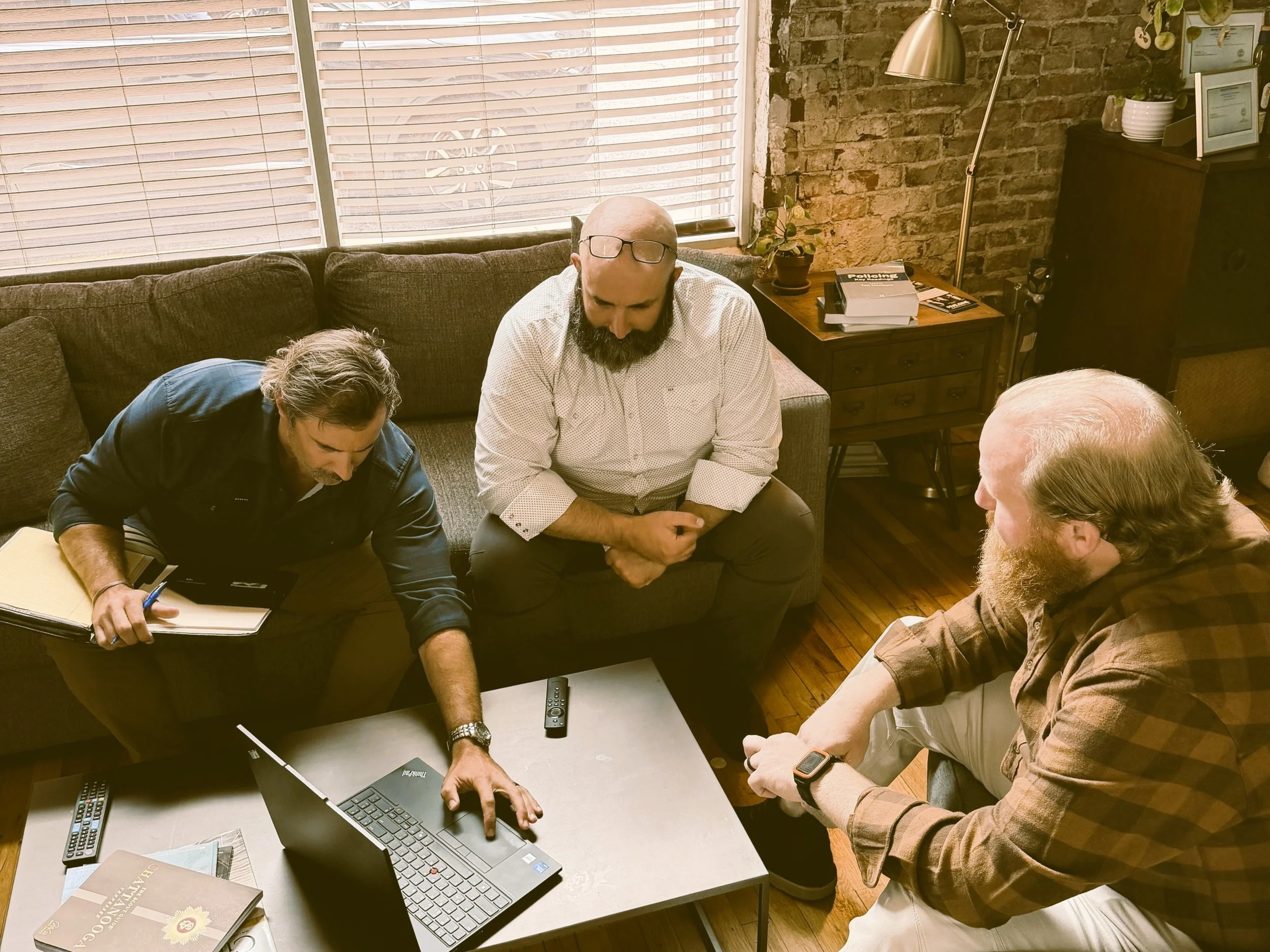 A group of four men engaged in a discussion around a coffee table with a laptop, remote controls, and magazines in a cozy, brick-walled room with a large window covered by blinds, a small table with books and a plant, and a tall floor lamp.