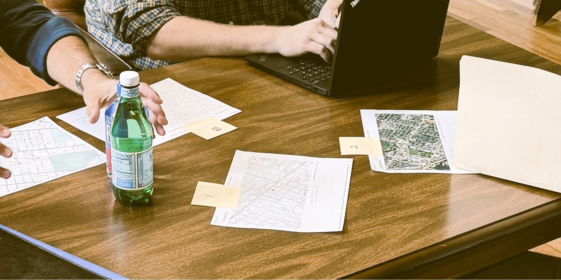People sitting at a wooden table with maps, papers, and a laptop, with one person reaching for a green bottled drink.