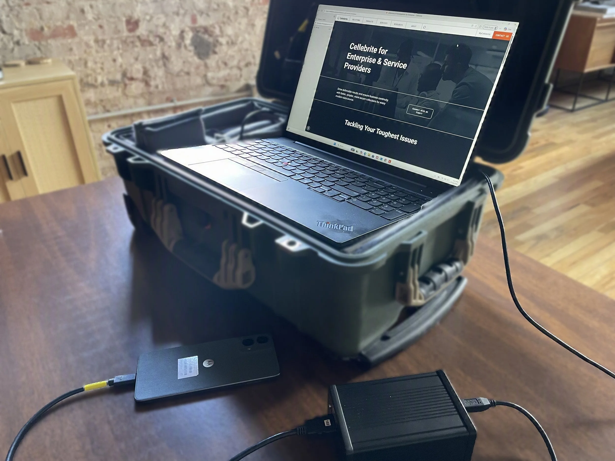 A laptop connected to a device inside a black protective case, with another laptop underneath, on a wooden table. The laptop screen displays a website called 'Cellebrite for Enterprise & Service Providers,' with a slogan that reads 'Tackling Your Toughest Issues.' The background features a brick wall and wooden furniture.