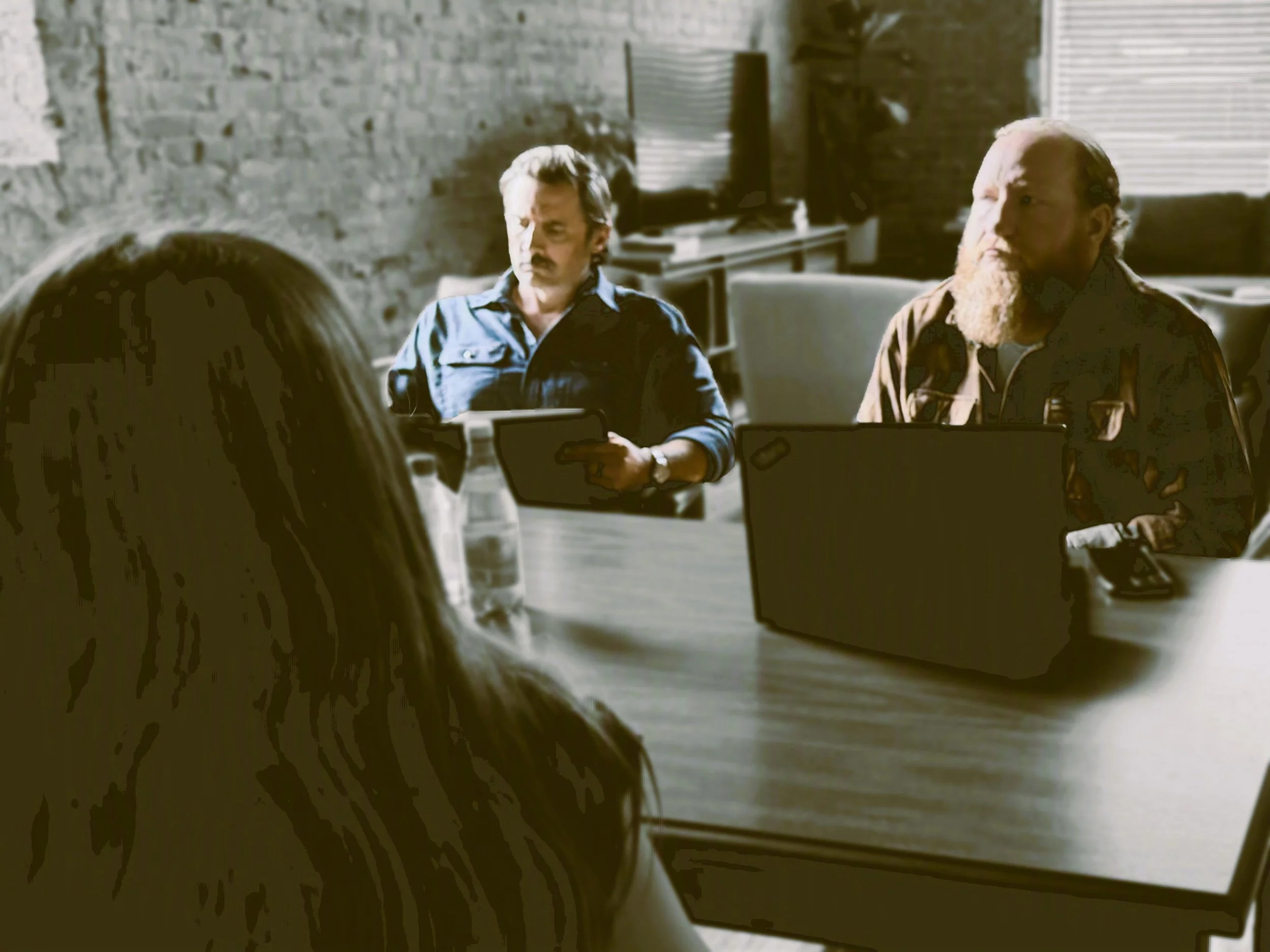 Two men with serious expressions sitting at a table in a modern office or meeting room with brick walls and a window with blinds, engaged in conversation.