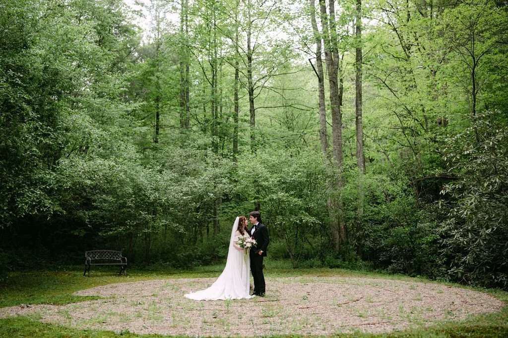 A bride and groom stand in a green forest clearing, holding hands and gazing at each other on their wedding day, with a bench in the background.