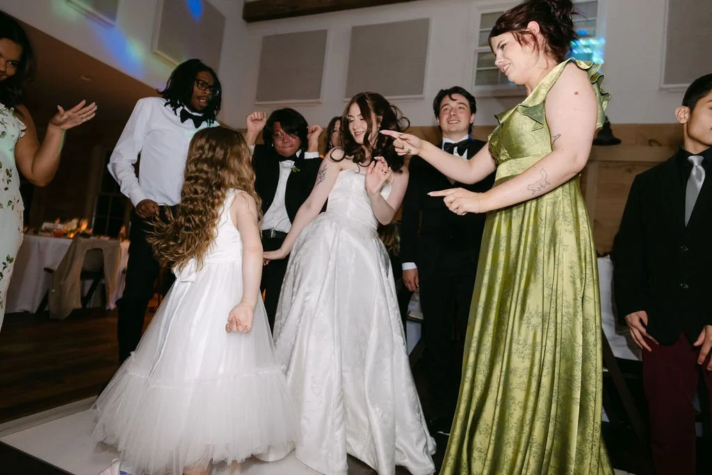 A group of people, including children in white dresses and adults in formalwear, dancing and enjoying a celebration at a wedding reception.