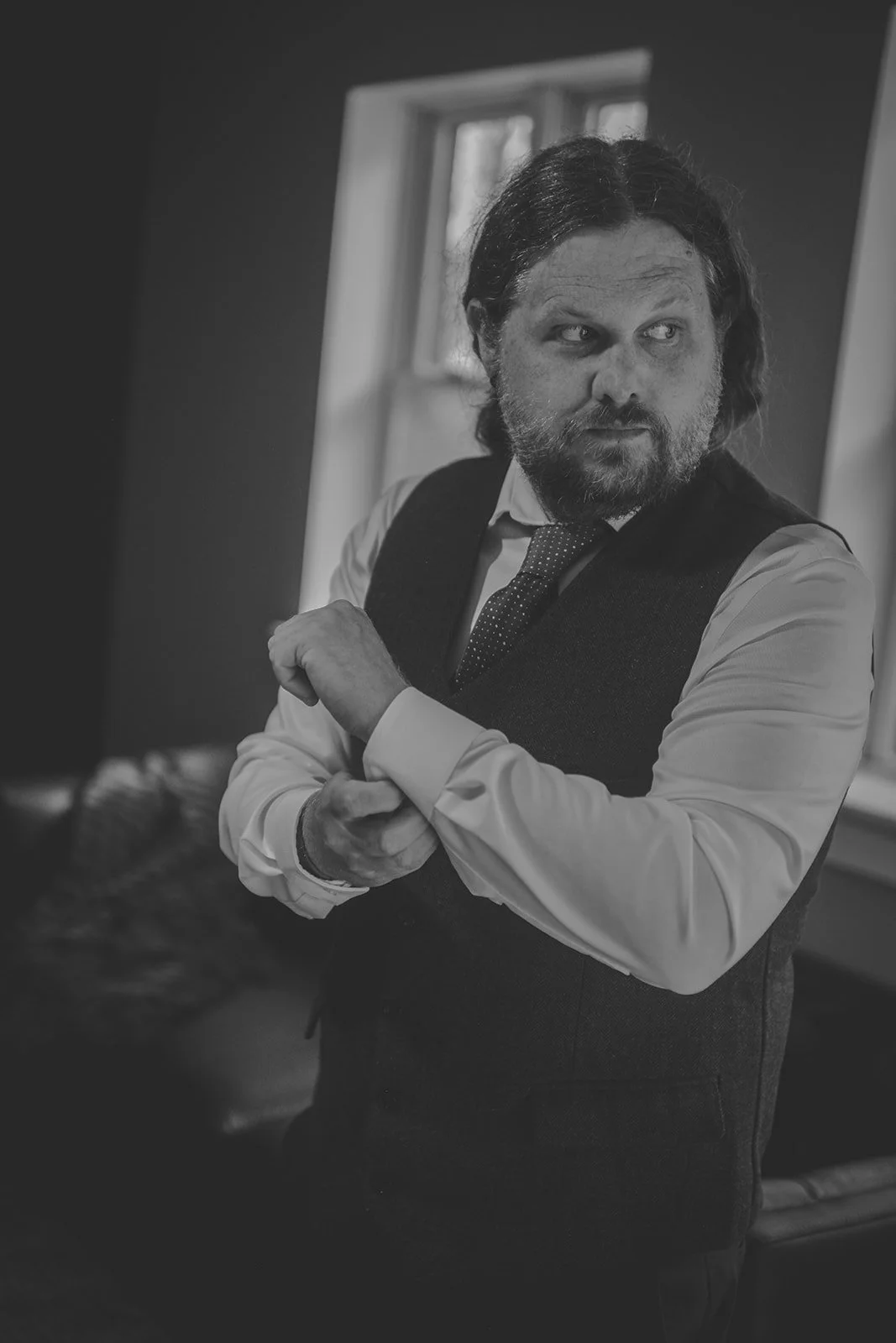 A man with shoulder-length hair and a beard, wearing a white shirt, a dark vest, and a tie, is adjusting his cufflink while looking to his right in a room with a window in the background.