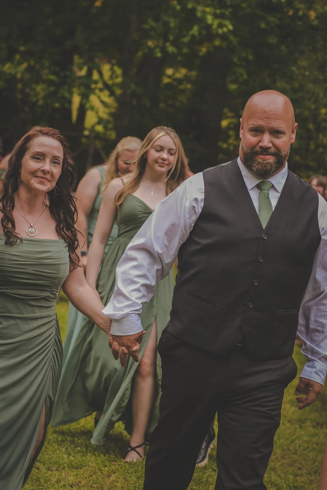 A group of people outdoors at a wedding or special event, holding hands and walking together on grass, with trees in the background.