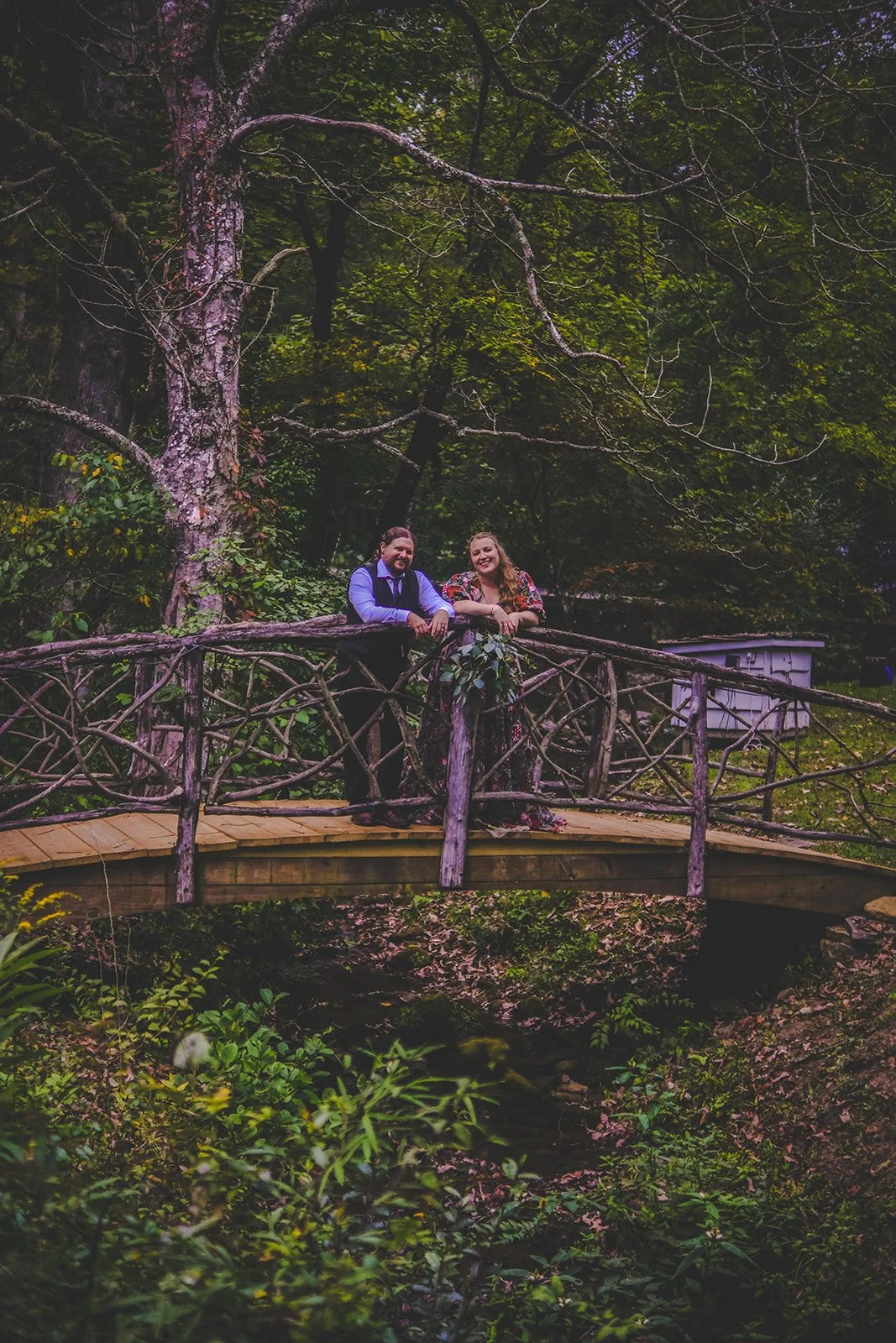 Two people, a man and a woman, standing on a wooden bridge in a lush, green forest, smiling and leaning on the rustic railing.