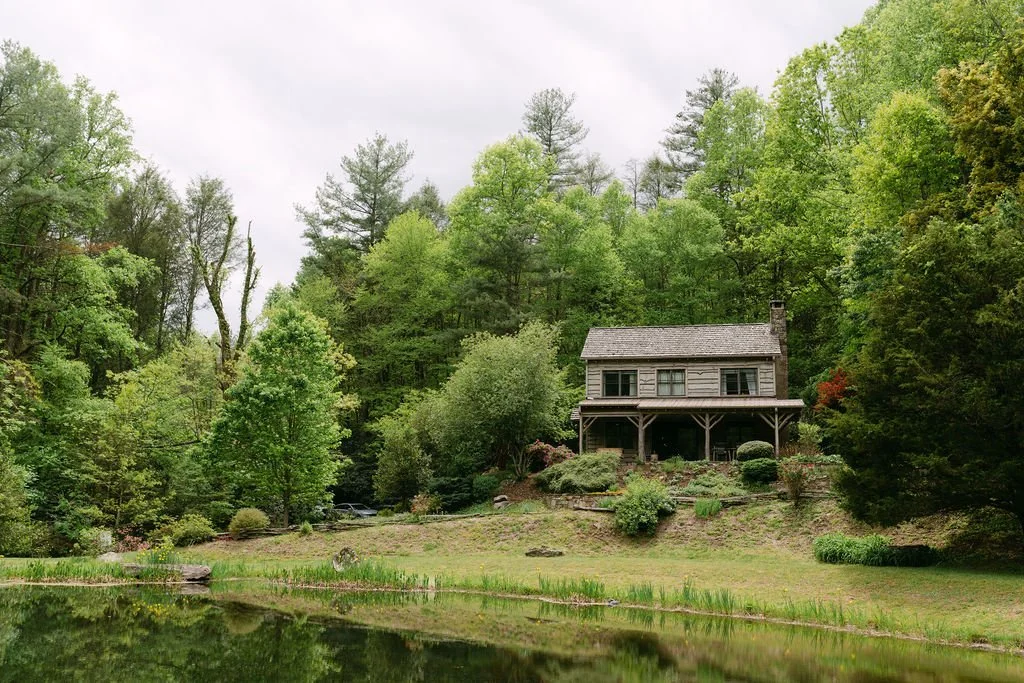 A rustic house on a hill surrounded by lush green trees and bushes, with a pond in the foreground reflecting the scene.