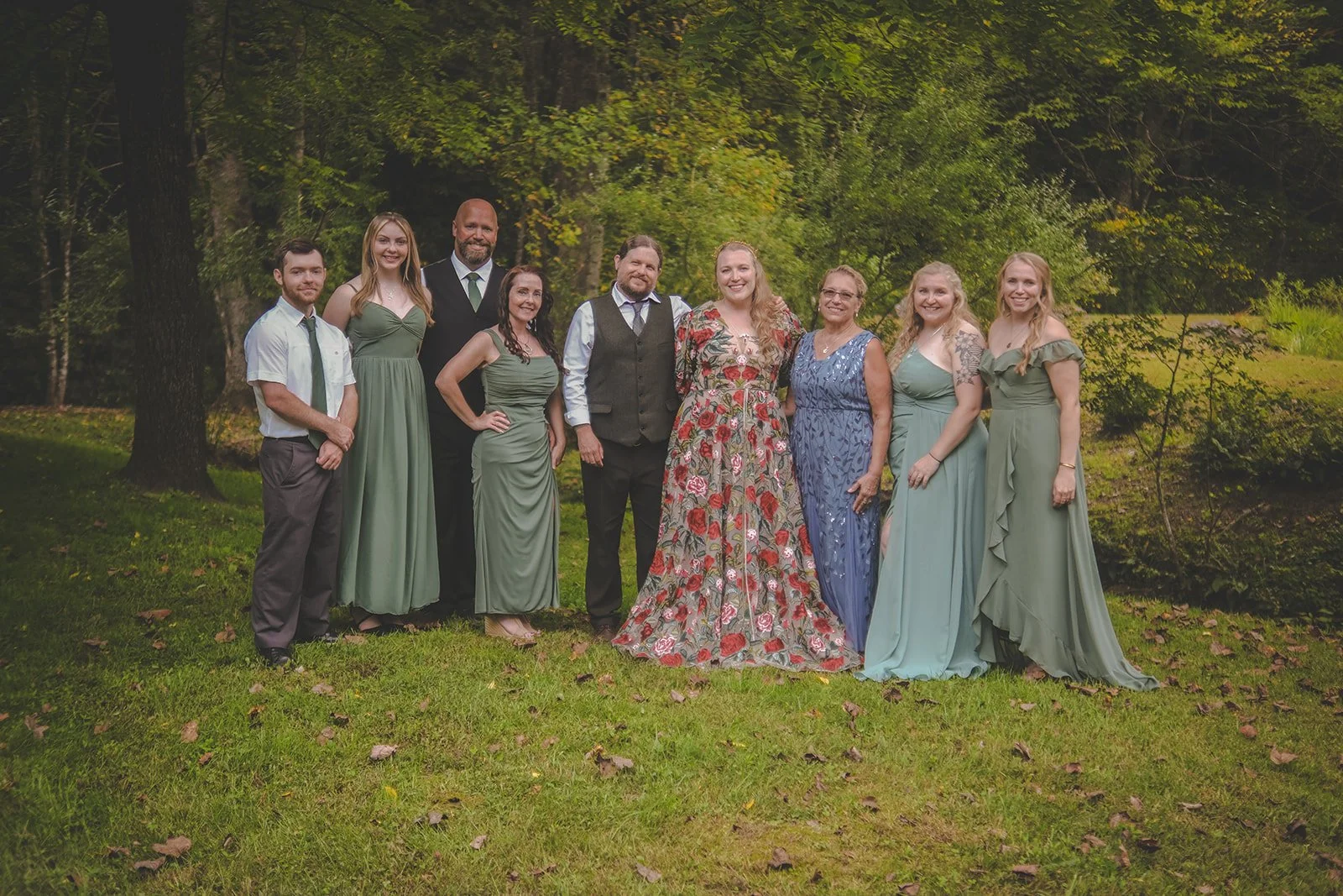 Group of nine people outdoors in a forested area, dressed in semi-formal attire.