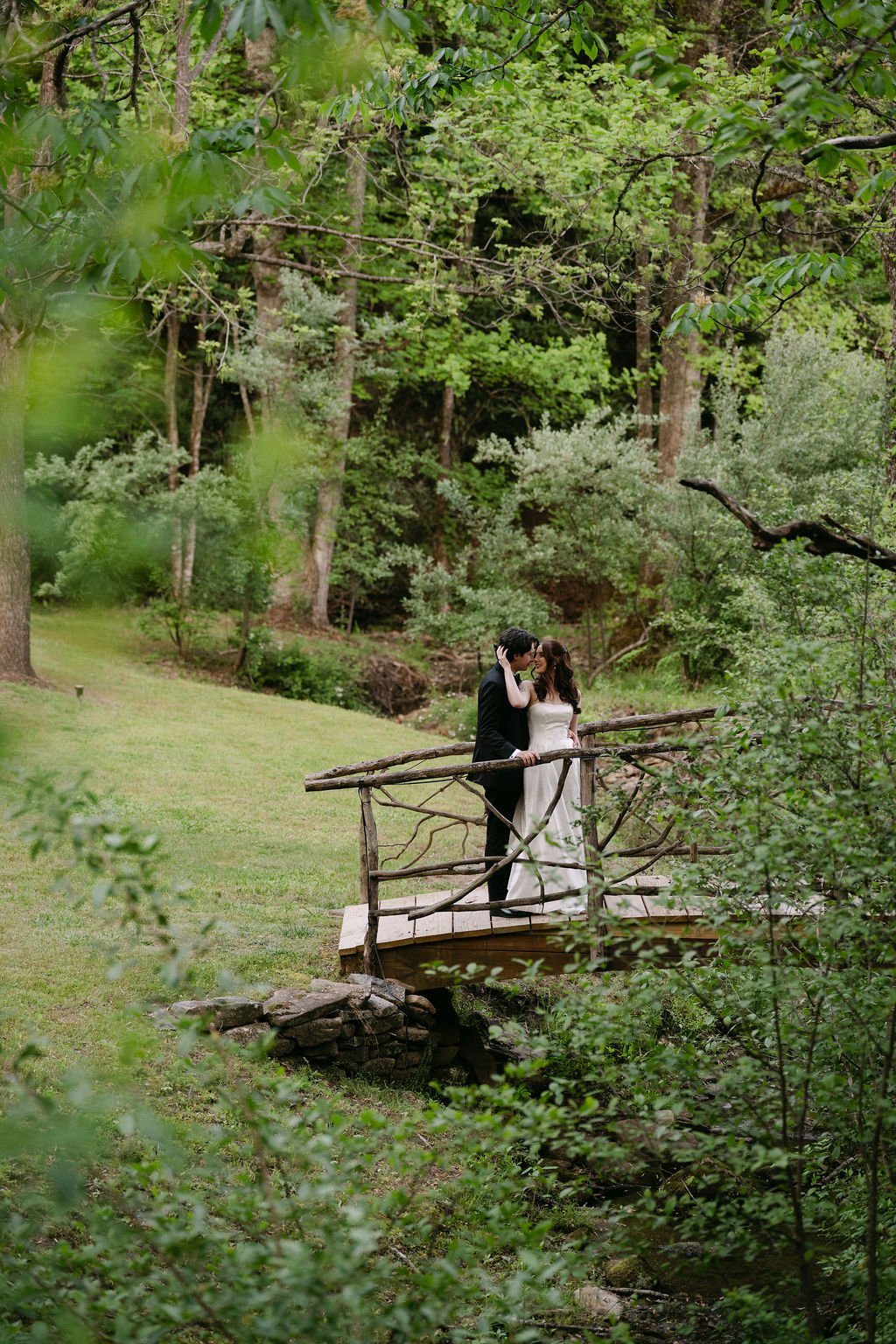 A bride and groom standing on a small wooden bridge in a lush green forest, sharing a tender moment.