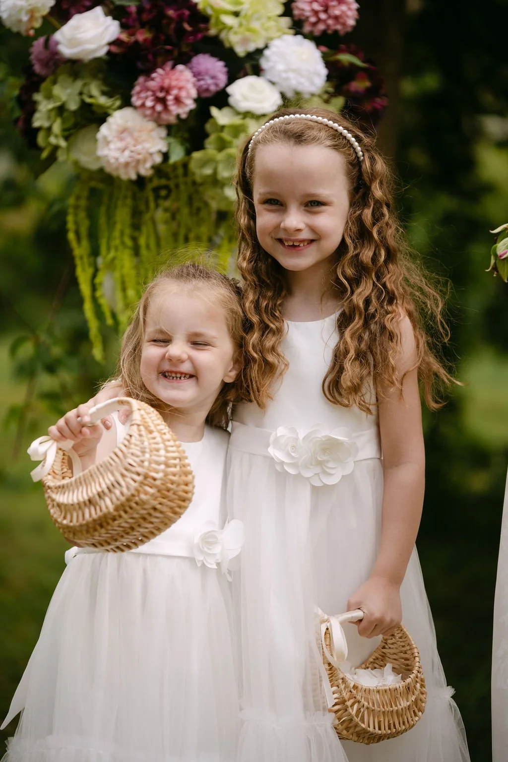 Two young girls in white dresses holding wicker baskets standing in front of hanging flowers at an outdoor event.