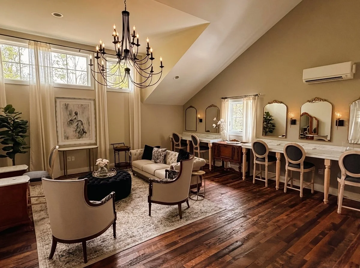 Living room with high ceiling, chandelier, cream-colored walls, multiple windows, and hardwood floors. It has a cream sofa, armchairs, a black tufted ottoman, and side tables. There is a vanity or desk area with mirrors and chairs, and wall-mounted l