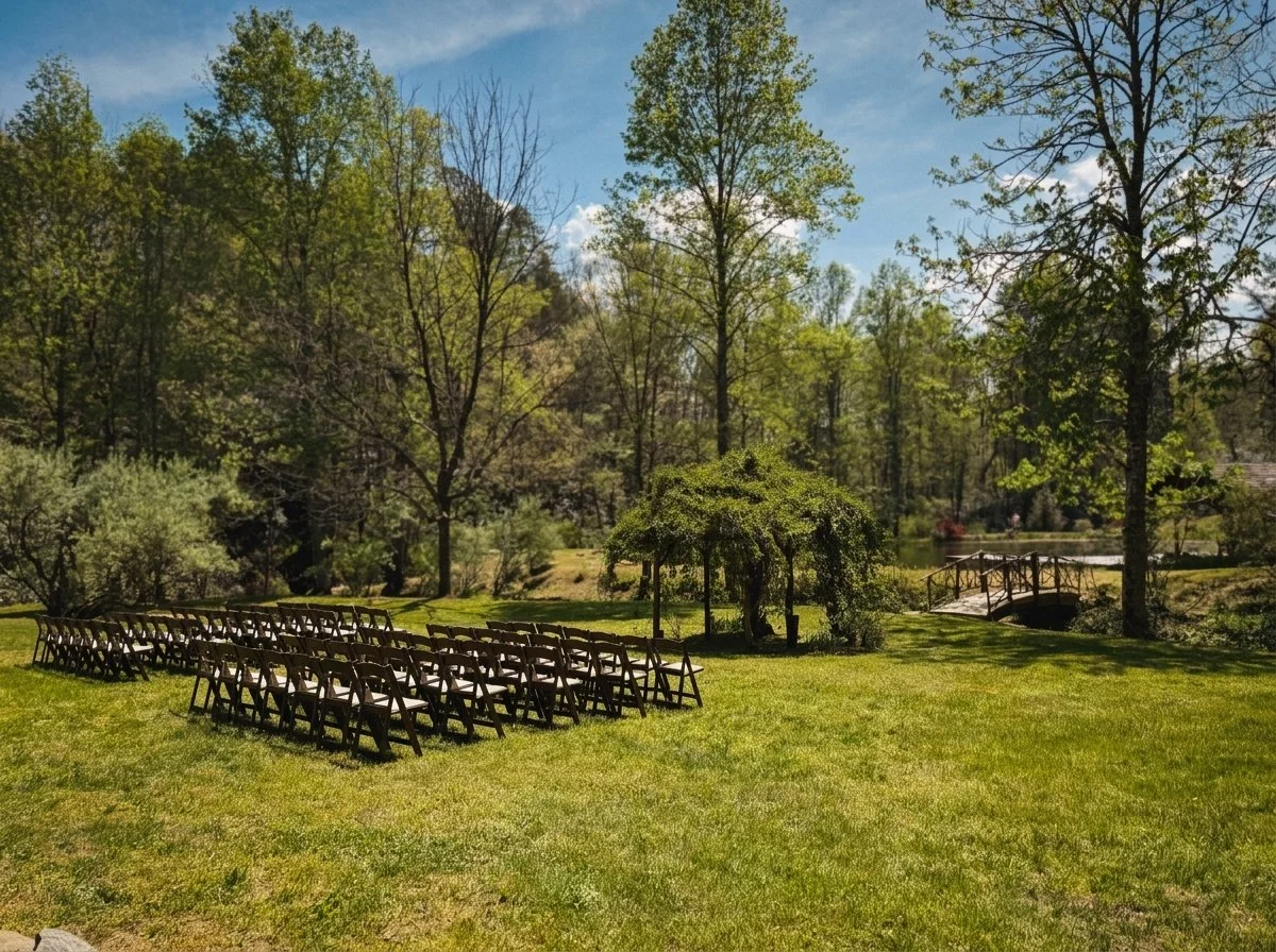 Chairs arranged in rows outdoors next to a small bridge over the water, surrounded by trees and greenery, on a sunny day.
