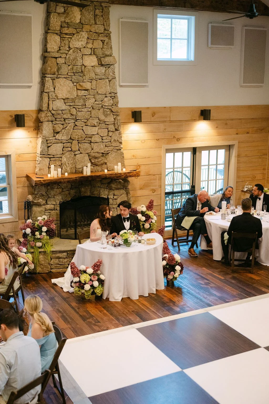 Wedding reception with a bride and groom seated at a decorated table near a stone fireplace, surrounded by guests in a rustic-style room with wood paneling, windows, and floral arrangements.
