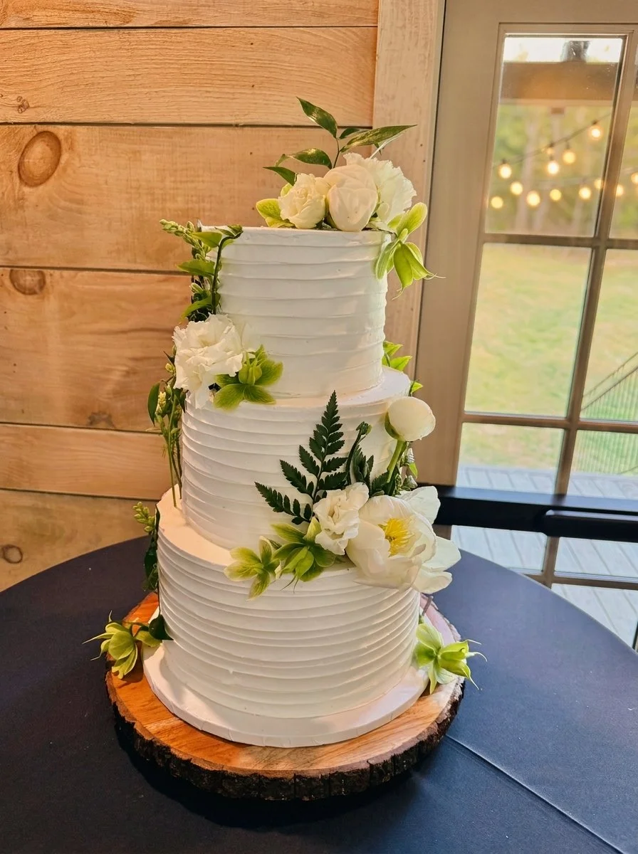 A three-tiered white wedding cake decorated with white flowers and green leaves, sitting on a wooden cake stand, with a window and string lights in the background.