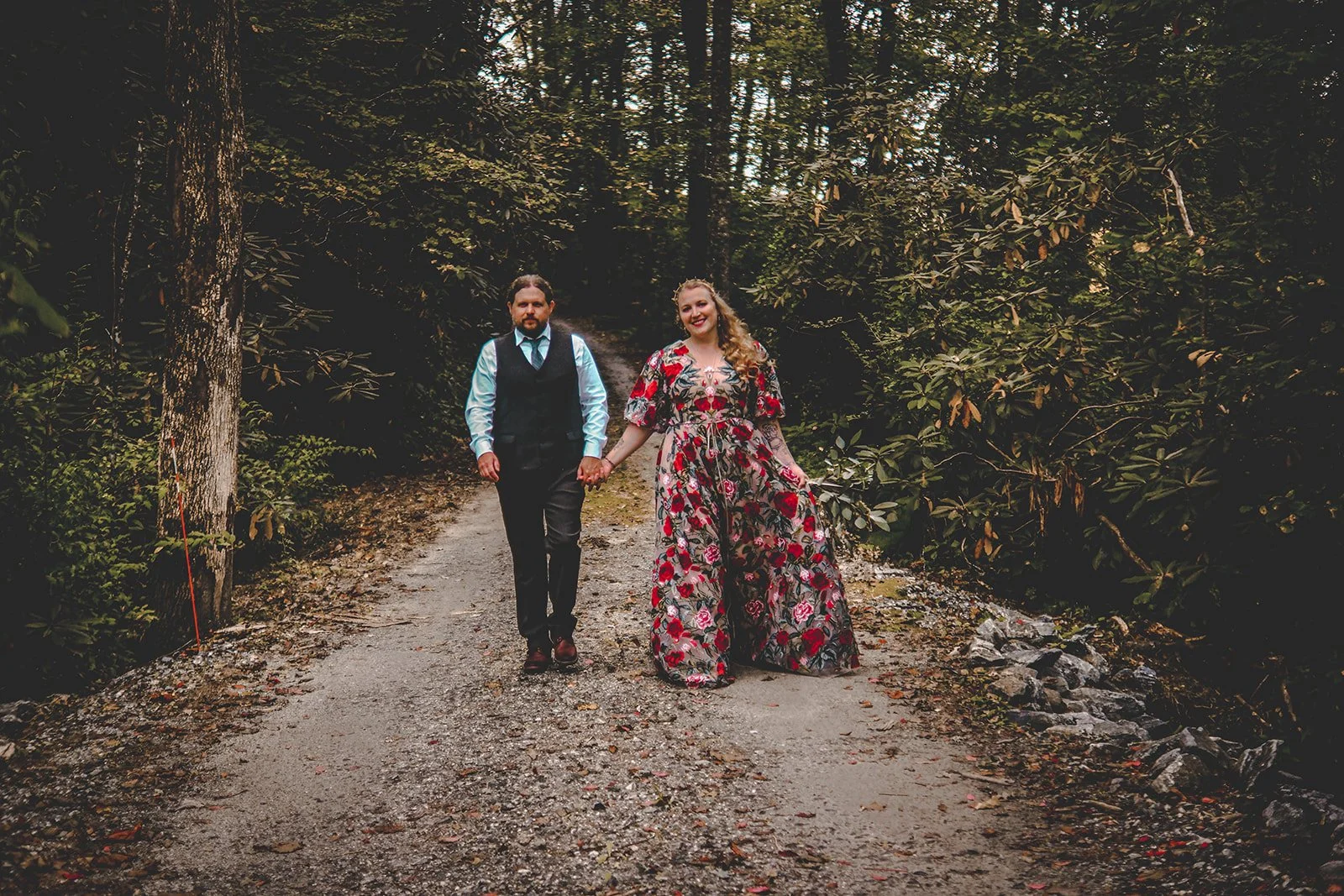 A couple holding hands and walking on a dirt path in a forest, with trees and greenery on both sides.