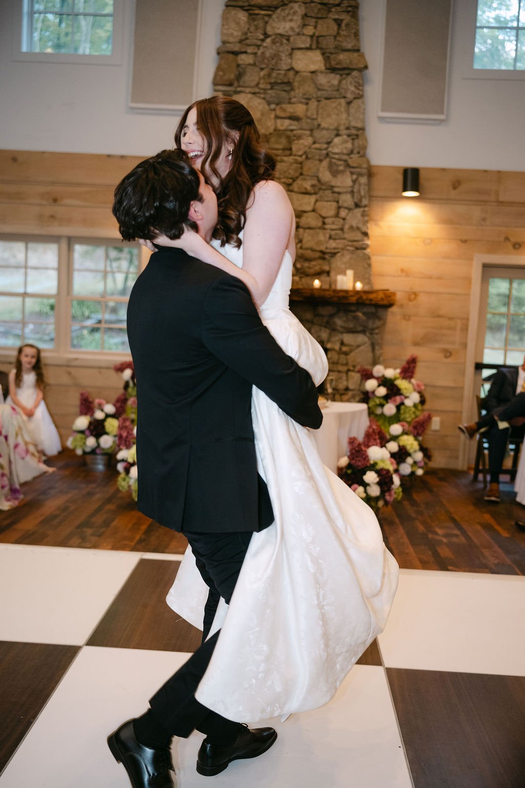 A groom in a black suit lifts a bride in a white wedding dress during their wedding dance in a rustic venue with wood walls, stone fireplace, and floral decorations.