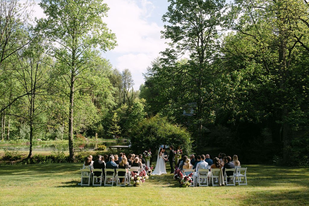 Outdoor wedding ceremony set in a lush, green garden with trees and a pond, where a bride and groom are exchanging vows in front of seated guests.