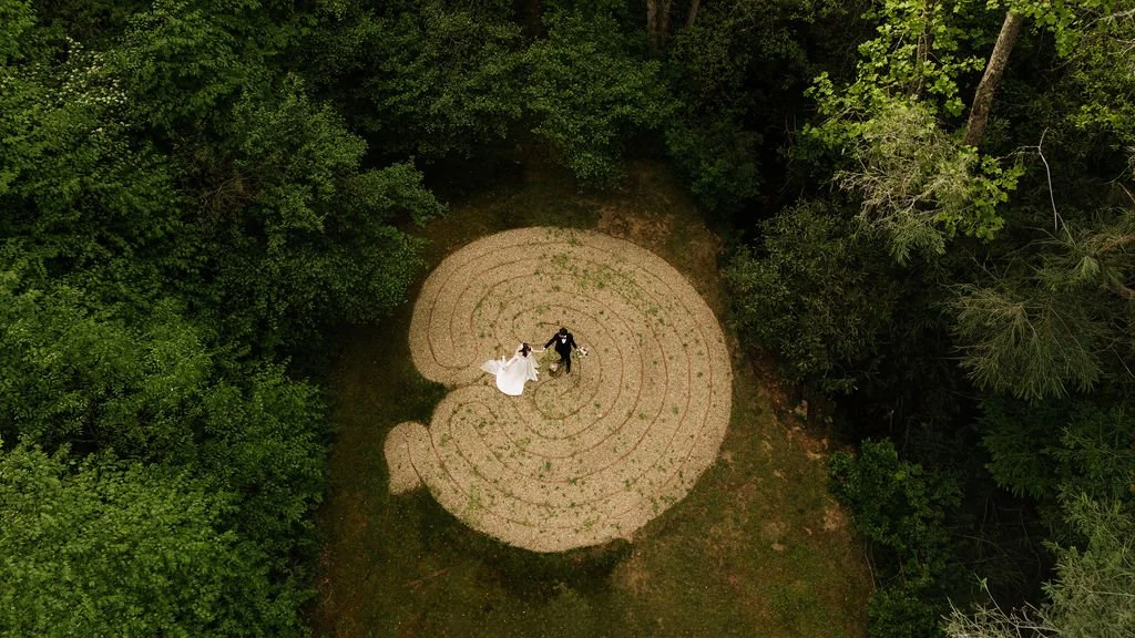 A couple in wedding attire dancing in a spiral-shaped clearing surrounded by green trees.