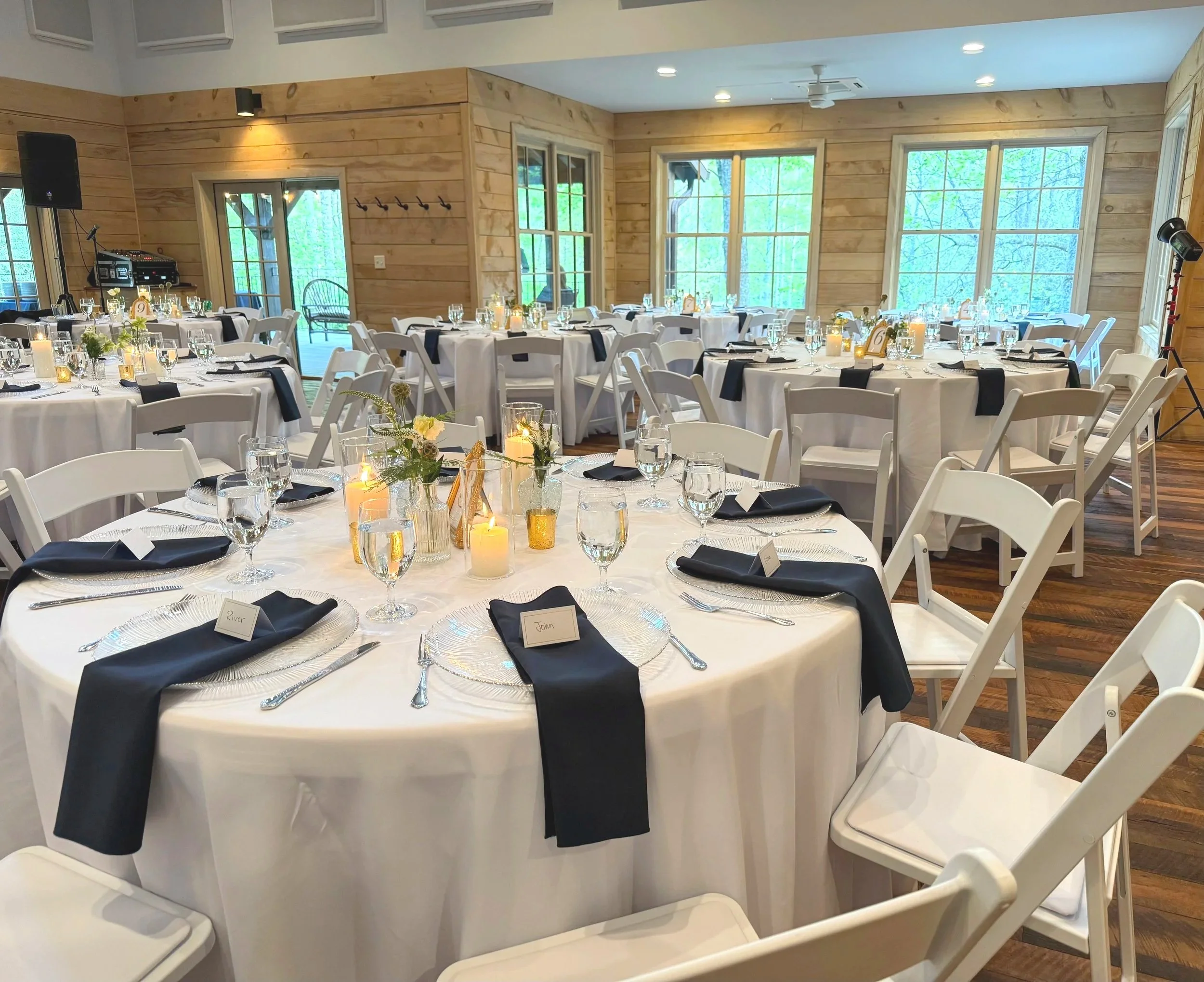Wedding or banquet reception with round tables covered in white tablecloths, decorated with candles and floral centerpieces, set with glasses, plates, cutlery, and black napkins. The room has wooden paneled walls, large windows showing greenery outside, and white folding chairs around the tables.