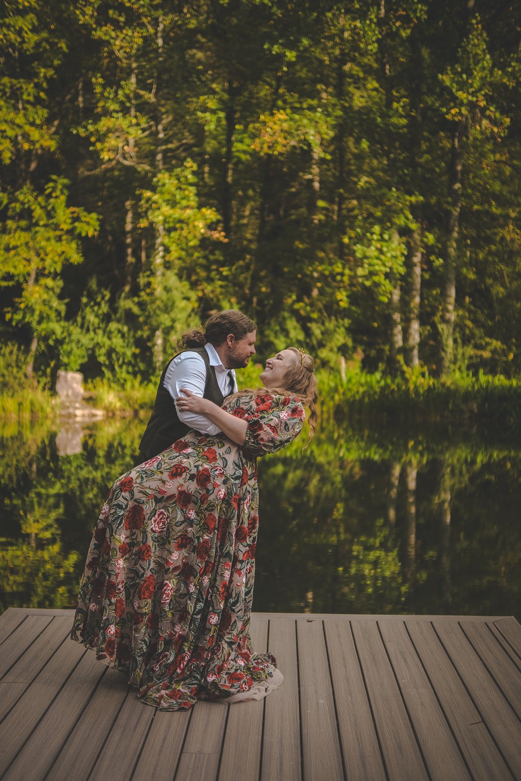 A man and woman, dressed in vintage clothing, share a romantic dance on a wooden dock by a lake, surrounded by lush green trees.
