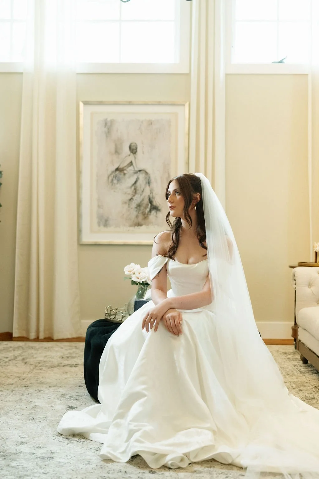 A bride sitting on a black ottoman in a well-lit room, wearing a white wedding gown and veil, with a vase of white roses beside her and a framed sketch of a woman in the background.