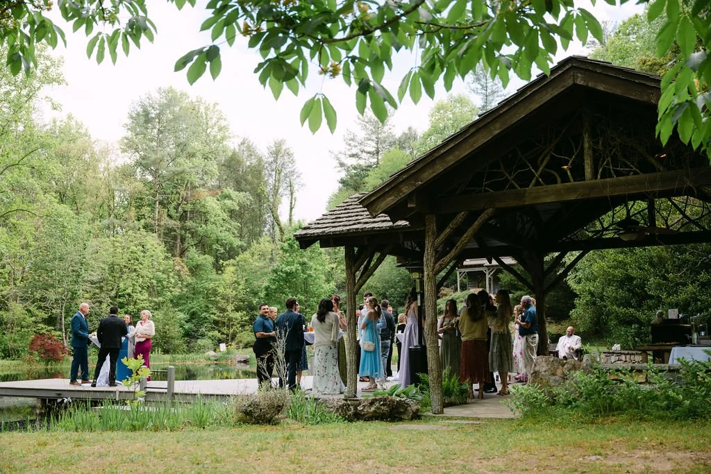 People gathered by a pond for a wedding ceremony in a lush, green outdoor setting, with a wooden pavilion on the right.