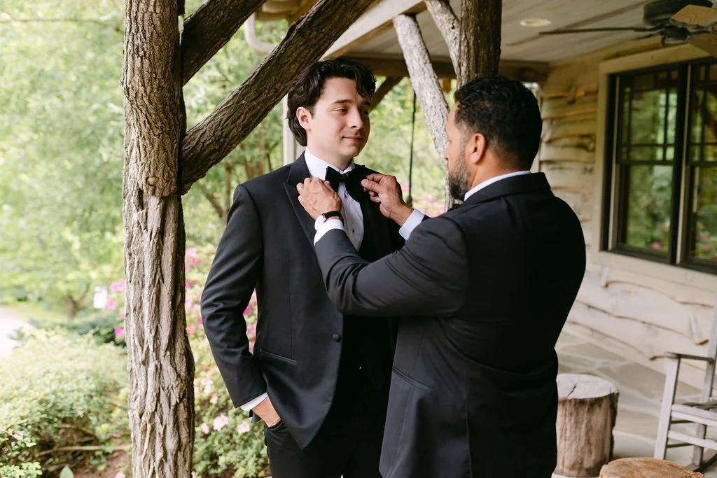 Two men in tuxedos, one helping the other adjust a bow tie outdoors next to a wooden house and trees.