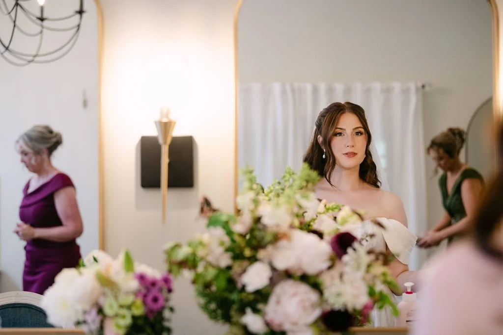 A bride with dark hair in a strapless white dress looking at a mirror, with floral arrangements in front of her and women preparing in the background.