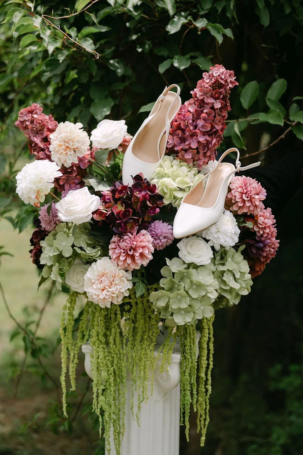 A floral arrangement on a white pedestal with pink, white, and purple flowers, including roses and hydrangeas, adorned with a pair of white high-heeled shoes.