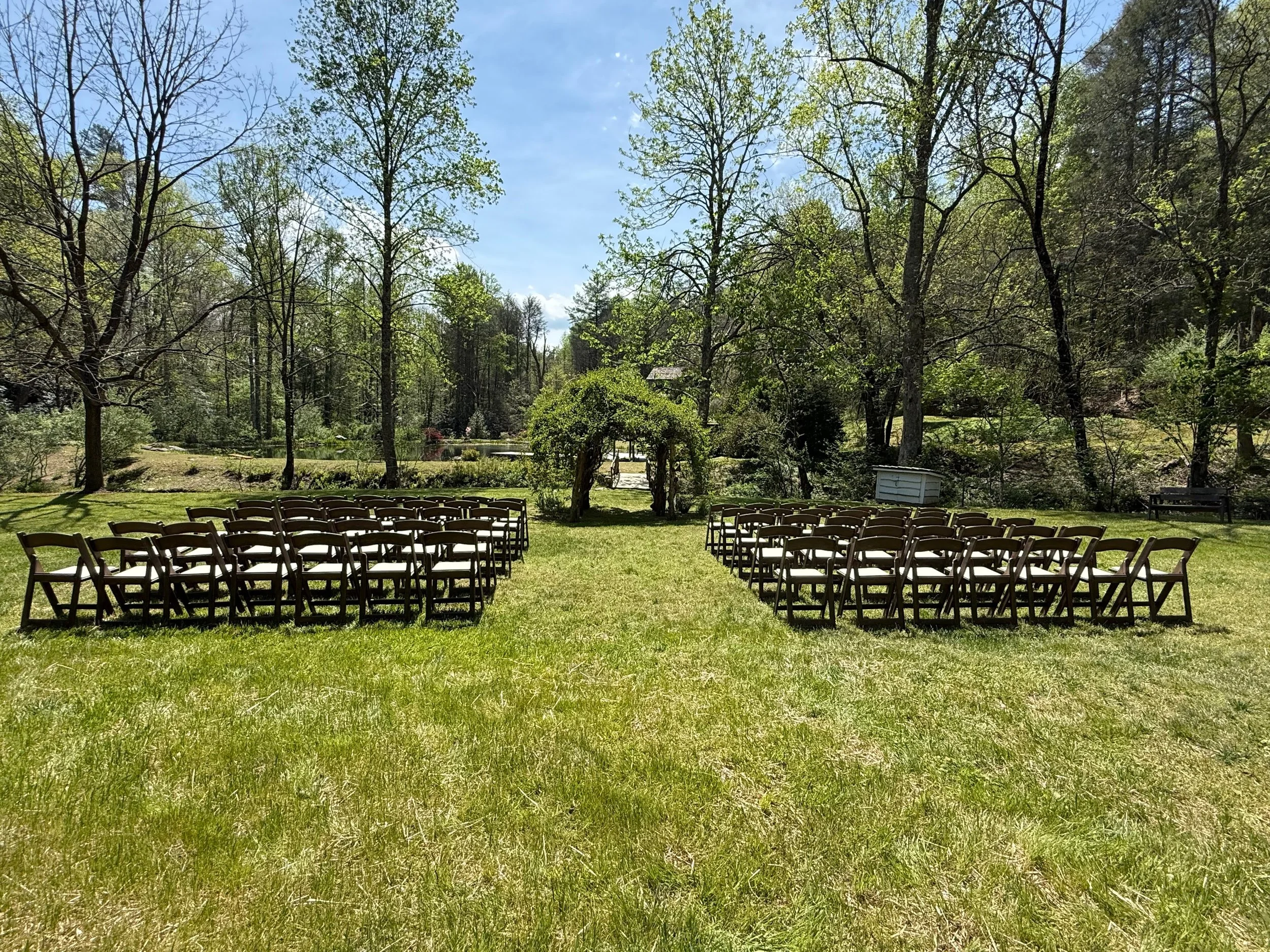 Outdoor wedding ceremony setup with rows of chairs on grass, archway, surrounded by trees and nature, under a blue sky.